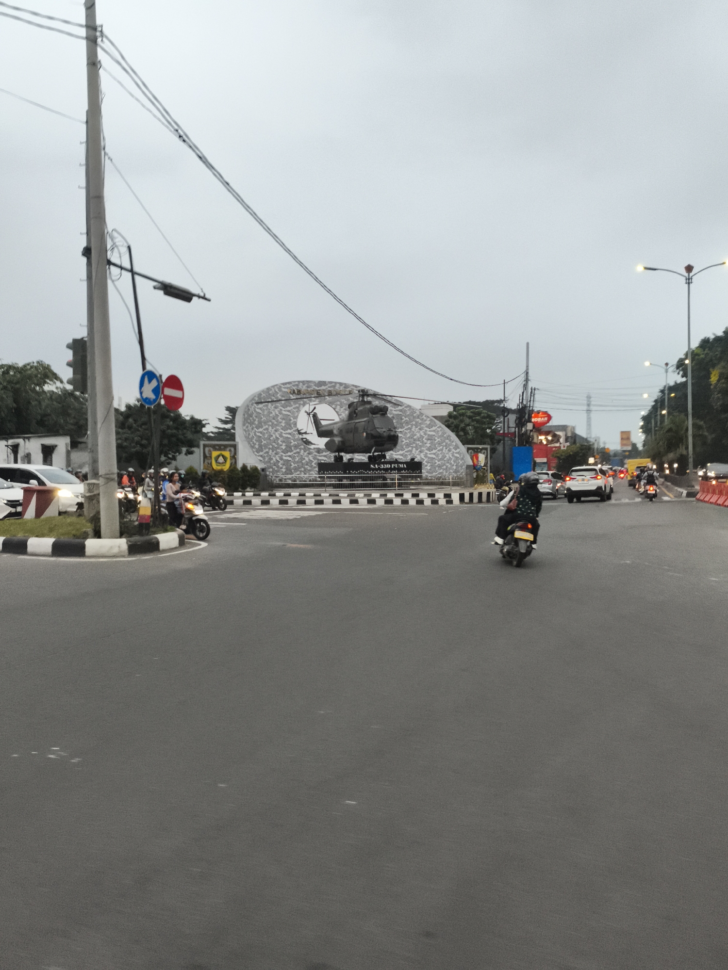 The image captures an urban street scene, likely a roundabout or a large intersection, under an overcast sky. The overall impression is that of a bustling city environment, consistent with Cibinong, Indonesia.
In the foreground, a wide asphalt road stretches towards the viewer. The midground is dominated by traffic; a person on a white scooter, wearing a dark jacket and a helmet, is seen from behind, riding through the intersection. To the left, several other people on motorcycles are stopped or waiting near a traffic island, alongside a white car with its headlights on. More vehicles, including cars and motorcycles, are visible further down the road to the right and left.
The central feature of the scene is a monument located in the middle of the roundabout. It showcases a dark-colored helicopter, appearing to be a full-scale replica or a real aircraft, mounted on a pedestal. Behind the helicopter is a large, curved, grey textured wall or structure. The monument is surrounded by a low, striped curb.
Notable details include illuminated streetlights, suggesting either a very cloudy day or the time being early morning or late afternoon/dusk. Power poles with a dense network of overhead wires crisscross the upper part of the image. Traffic islands are marked with black and white stripes, and red and white plastic barriers line the right side of the road. Two distinct traffic signs, one blue (possibly indicating direction or allowed vehicles) and one red (likely a prohibitory sign like 'no entry'), are visible on the left side of the road.
Visible text includes "SA-330 PUMA" on the base of the helicopter monument, identifying the aircraft model. Above it, the word "NARASOMA" is also visible on the monument's background structure. In the right background, a red sign with the text "ROBAN" can be seen.