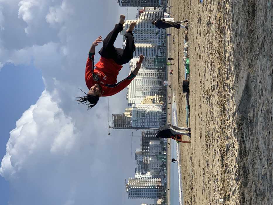 A man with dreadlocks wearing a red jacket and black pants is mid-air performing a backflip on a beach. Behind him, there are several people, buildings under construction, and finished buildings. The beach is sandy, and the sky is partly cloudy. Another person is bending over in the lower right of the photo.