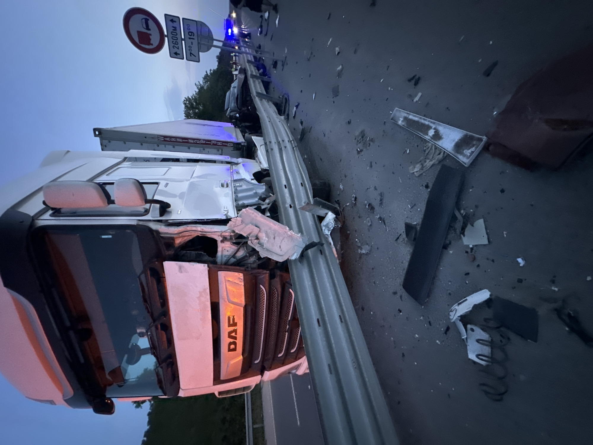 The image depicts the aftermath of a multi-vehicle collision on a highway in Chirpan, Bulgaria, observed at dusk or nighttime. A white DAF semi-truck is positioned prominently in the foreground, facing the camera at an angle. Its front end exhibits severe damage, with the bumper, grille, and headlights crushed and fragmented. The DAF logo on the grille is still visible amidst the deformation. The truck has impacted and extensively damaged a metal guardrail, which is bent inwards and broken at the point of impact.
Scattered on the asphalt road surface around the truck and guardrail are numerous pieces of vehicle debris, including plastic fragments, metal components, and a coiled spring. Further down the road, behind the damaged semi-truck, a dark-colored passenger vehicle is visible, also appearing to be damaged. Blue flashing lights, consistent with emergency services (police or ambulance), are discernible in the distance, illuminating the scene. Several individuals are present in the background near the emergency lights and the damaged passenger vehicle, though their specific actions are not discernible.
Two road signs are visible on the right side of the road. The upper sign is a circular red and white regulatory sign depicting a truck and a car, indicating a restriction, possibly "no overtaking for trucks." Below it, a rectangular white sign displays "↑ 2600M ↑" and "7:00 – 19:00", suggesting a time-based restriction (between 7 AM and 7 PM) applicable over a distance of 2600 meters. The sky in the background is a deep twilight blue, and a line of trees is visible along the roadside.