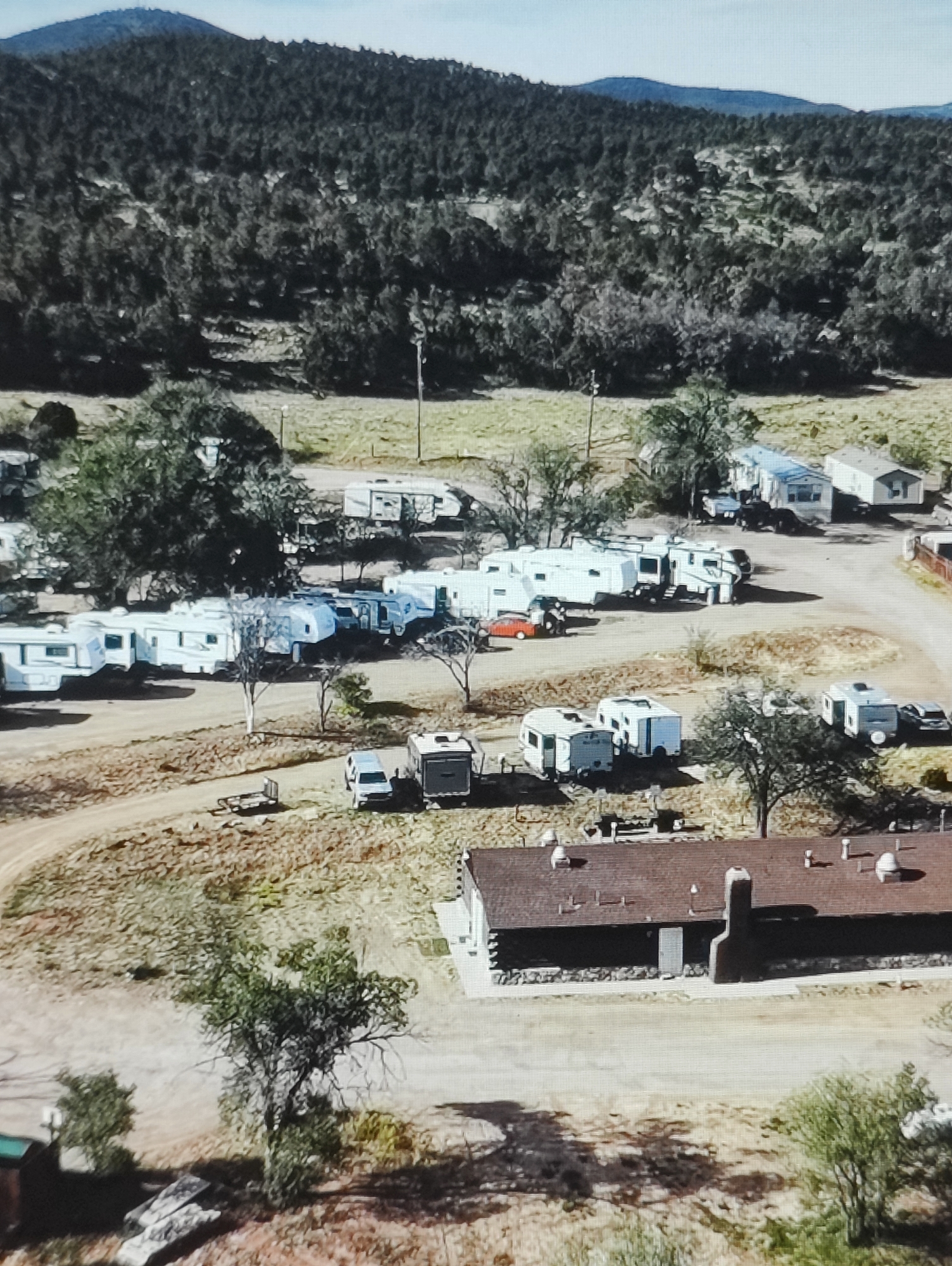 The image shows an aerial view of a mobile home park or RV campground nestled in a hilly, wooded area. Numerous white RVs and trailers of various sizes are parked in rows on dirt roads. A few smaller, white campervans or travel trailers are also visible. In the foreground, a low-slung building with a dark, pitched roof, likely a communal facility or office, is situated. The landscape is characterized by dry, scrubby vegetation, scattered trees, and a distant, hazy mountain range under a pale sky. There are no people clearly visible in the image. The scene appears to be a rural or semi-rural setting. The location is not in Yarmolyntsi, Ukraine, as the landscape and architecture do not match that region.