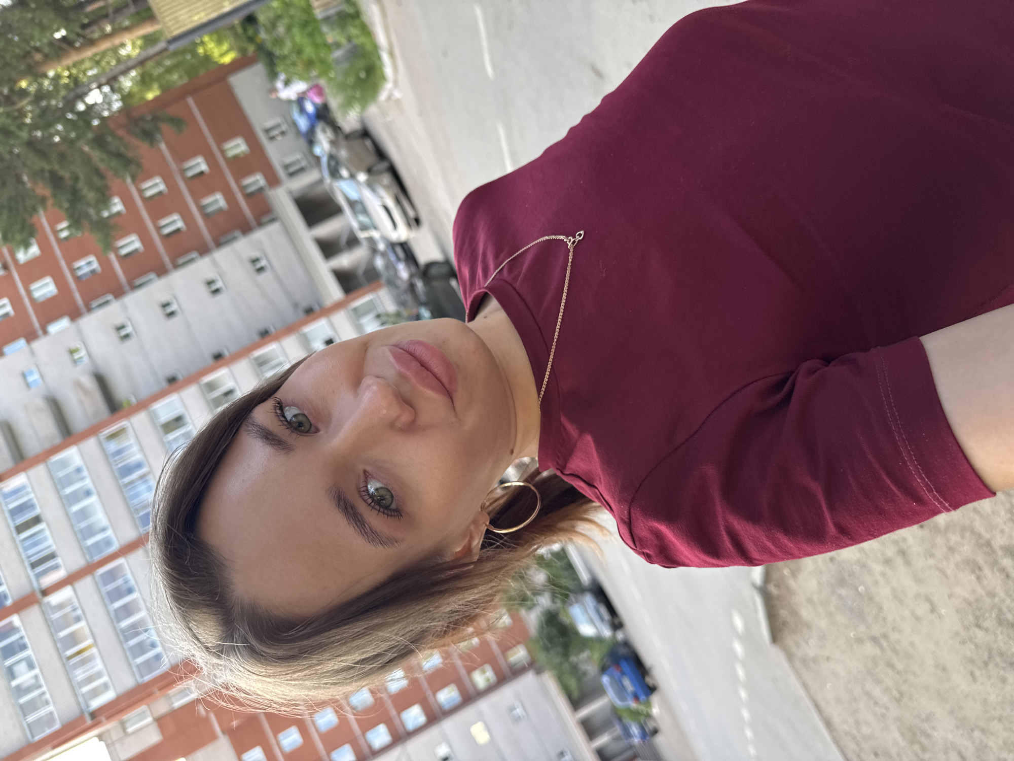 In Chaikovsky, Russia, a young woman with light brown hair and green eyes takes a selfie outdoors. She is wearing a maroon t-shirt and a gold necklace with a pendant. Her hair is pulled back in a ponytail, and she has large hoop earrings. The background shows a tall apartment building with red and grey brickwork and multiple windows. Several cars are parked in the street and on a sidewalk. Green trees and foliage are also visible.
