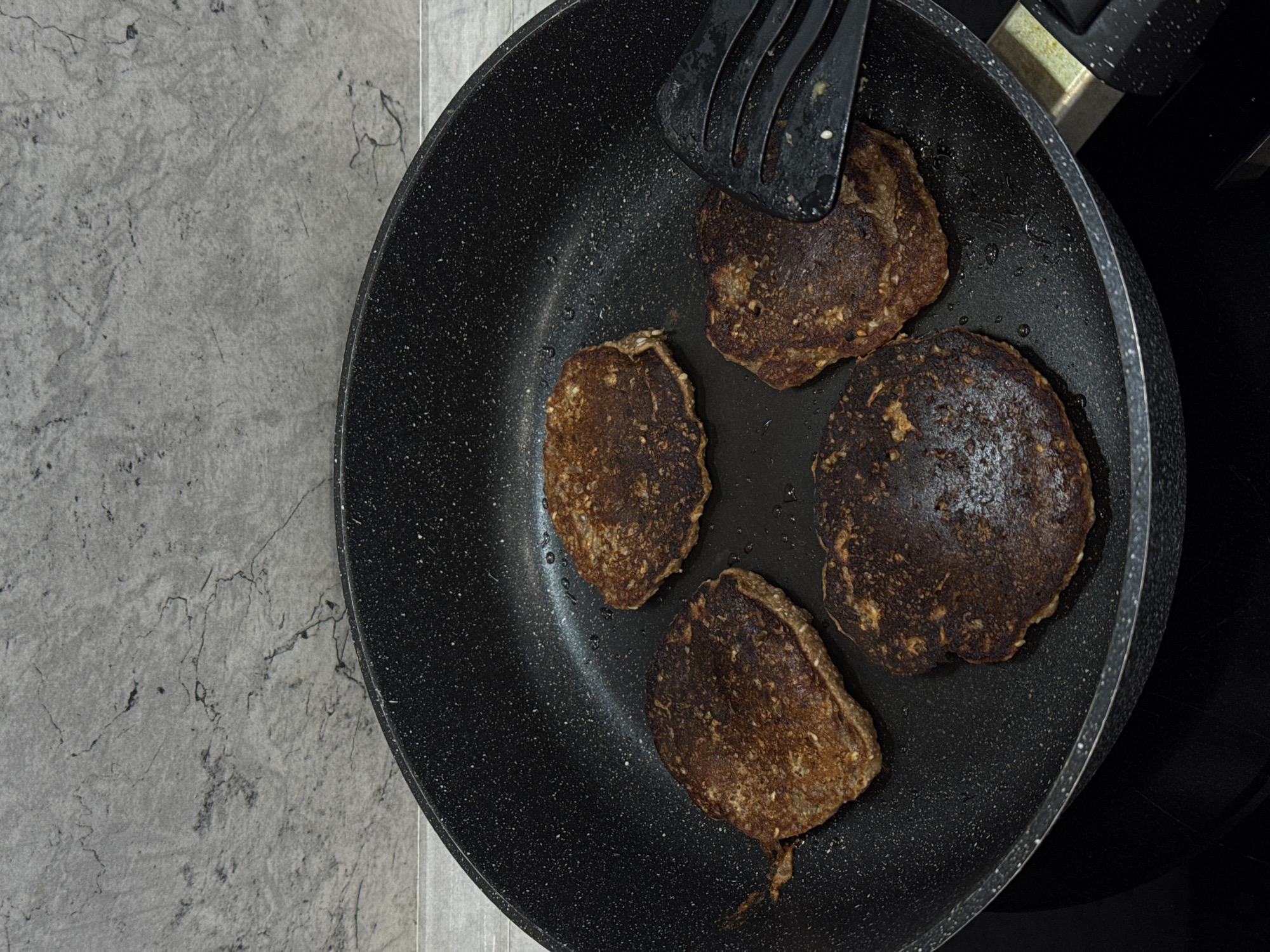 A high-angle shot shows a black frying pan containing four browned pancakes on a gray countertop, with a spatula resting on one pancake. The location is in an Unknown city, Unknown.