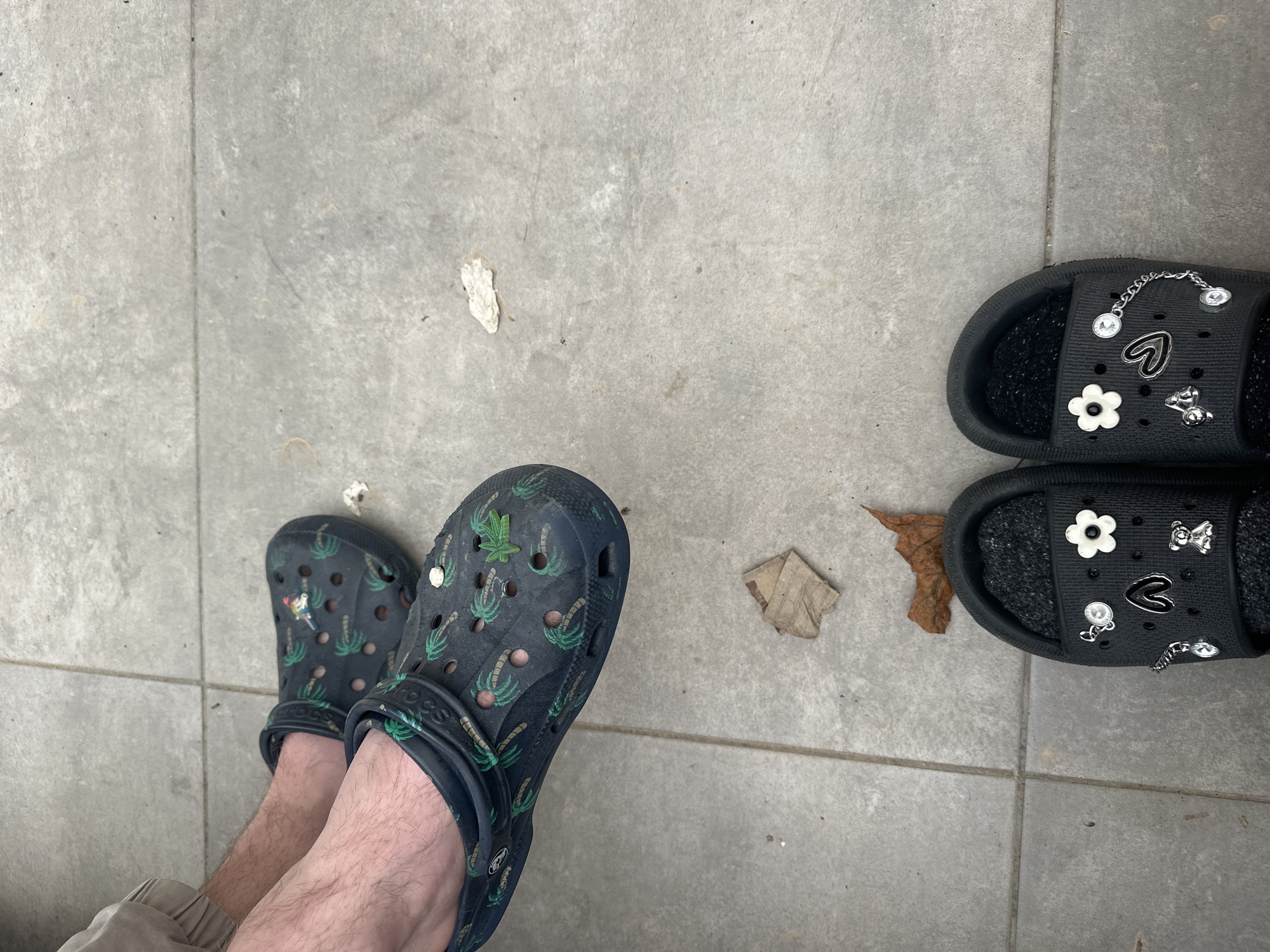 The image shows a view of the ground. A pair of feet wearing patterned Crocs are visible in the lower left. To the right are a pair of black sandals decorated with white flowers, hearts and other charms. The ground is made of gray tiles. There are small pieces of organic matter (leaves, etc.) scattered on the tiles. The location is unspecified, and Ramat Gan, Israel, is not indicated.