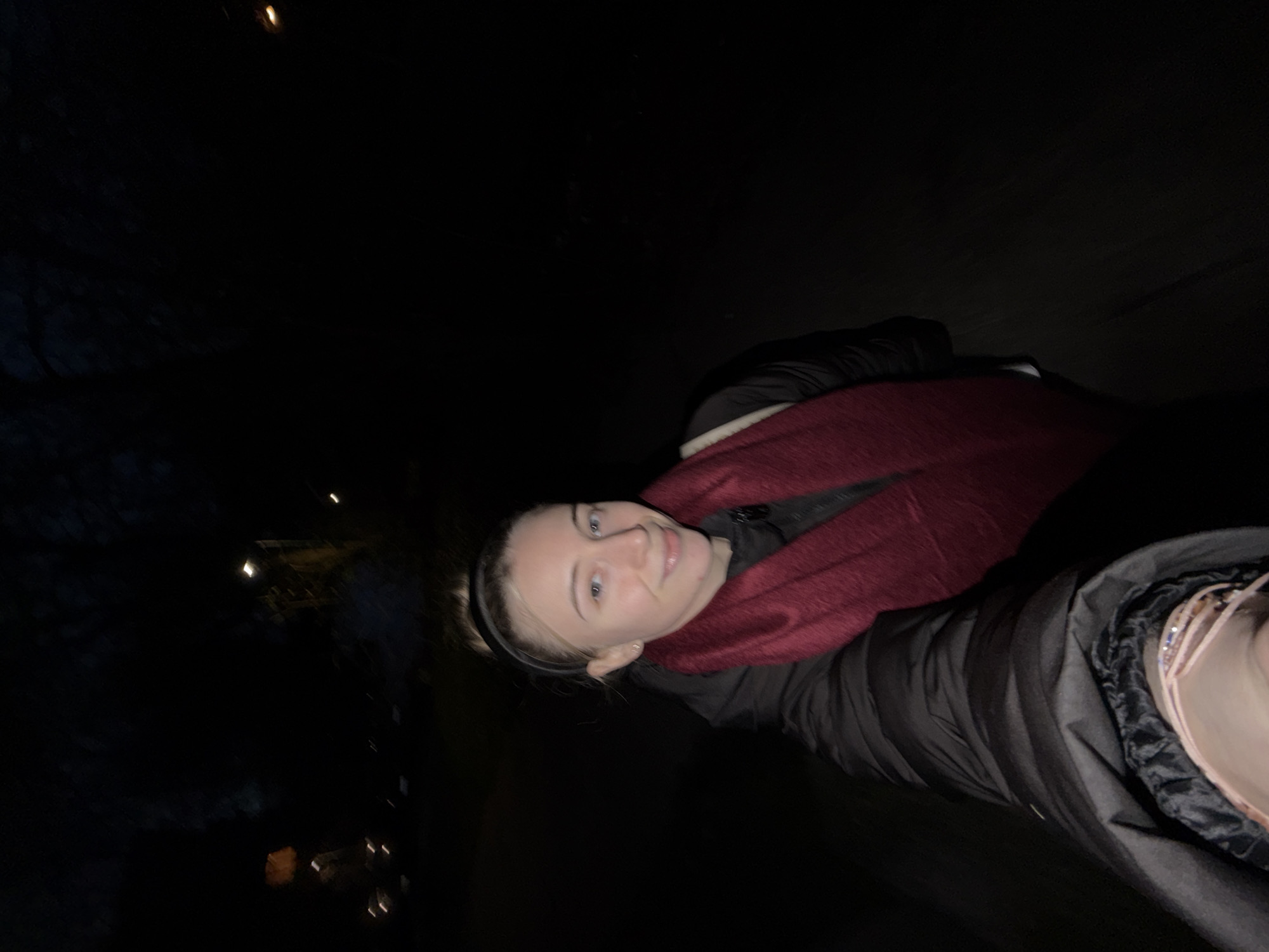 A female is photographed in an outdoor setting at night. The scene is dimly lit, with light sources coming from the left side of the frame, possibly from streetlights. The female is wearing a dark-colored, puffy jacket with a red scarf wrapped around her neck. She also has a black headband. The background is dark, with possible trees and building lights. The location cannot be confirmed as Plovdiv, Bulgaria.