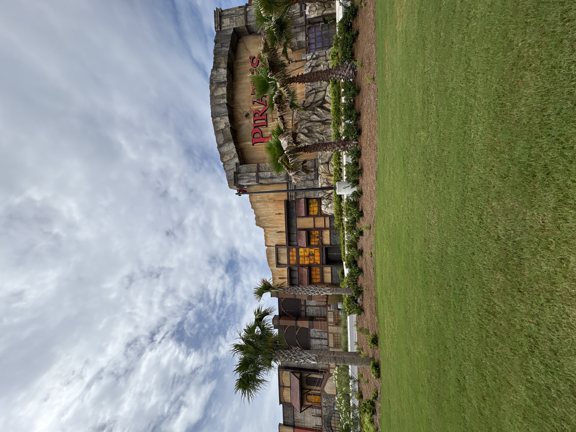 The image captures the "Pirates" building in Panama City Beach, United States. The building has a theatrical, aged appearance, featuring stone-like construction, weathered wooden elements, and a large, arched entrance with the word "PIRATES" in red lettering. Palm trees are positioned in front of the building. A green lawn stretches out beside the building. The sky above is partly cloudy.