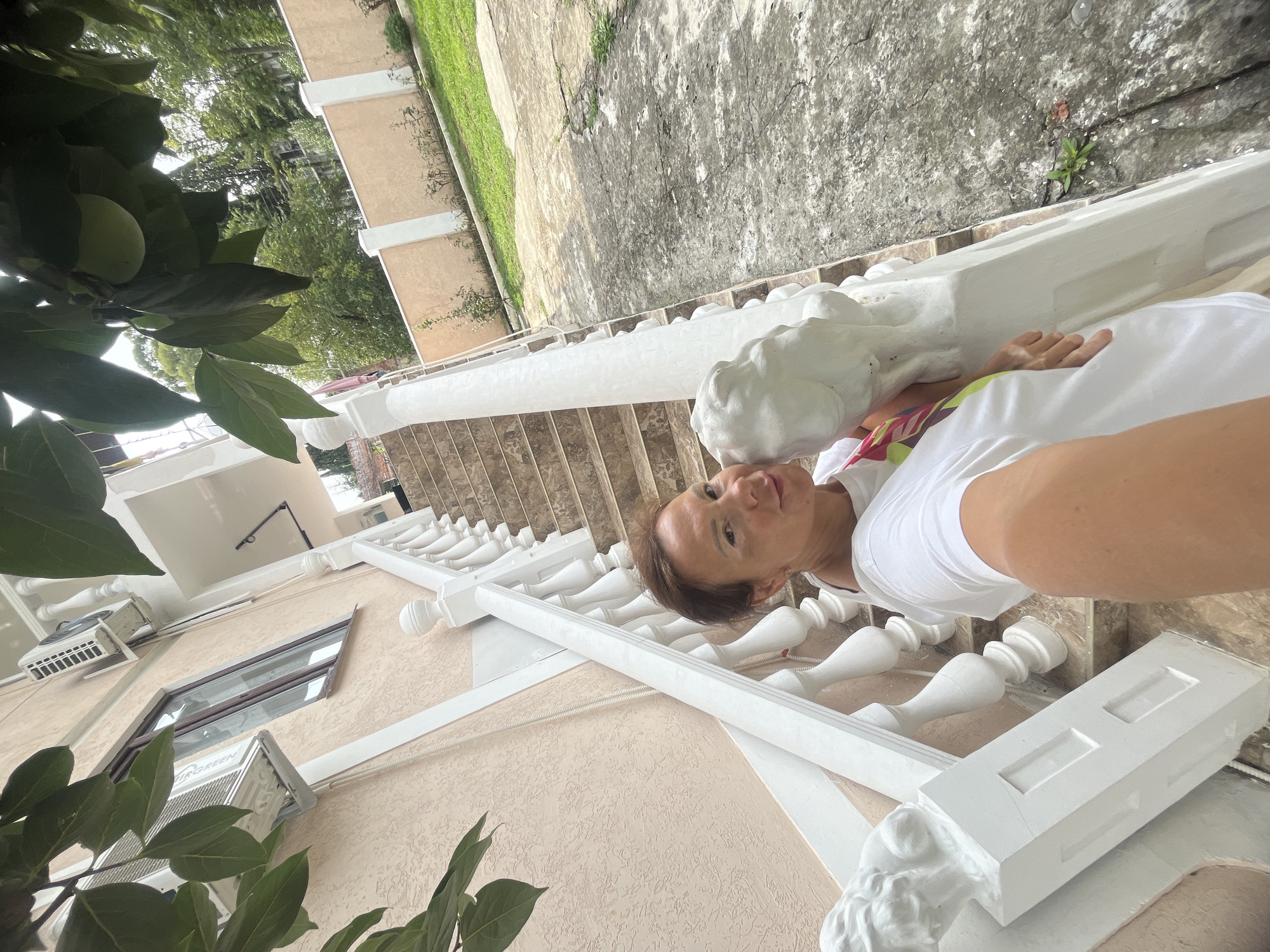 A woman takes a selfie while standing on a concrete path next to a white staircase with ornate balusters. She is wearing a white t-shirt and has her arm extended to take the photo. The staircase leads up to a pale pink building with an air conditioning unit visible on the exterior wall. In the background, there is a green lawn, a light brown fence, and trees. The scene appears to be outdoors in a residential area of Sokhumi, Abkhazia.
