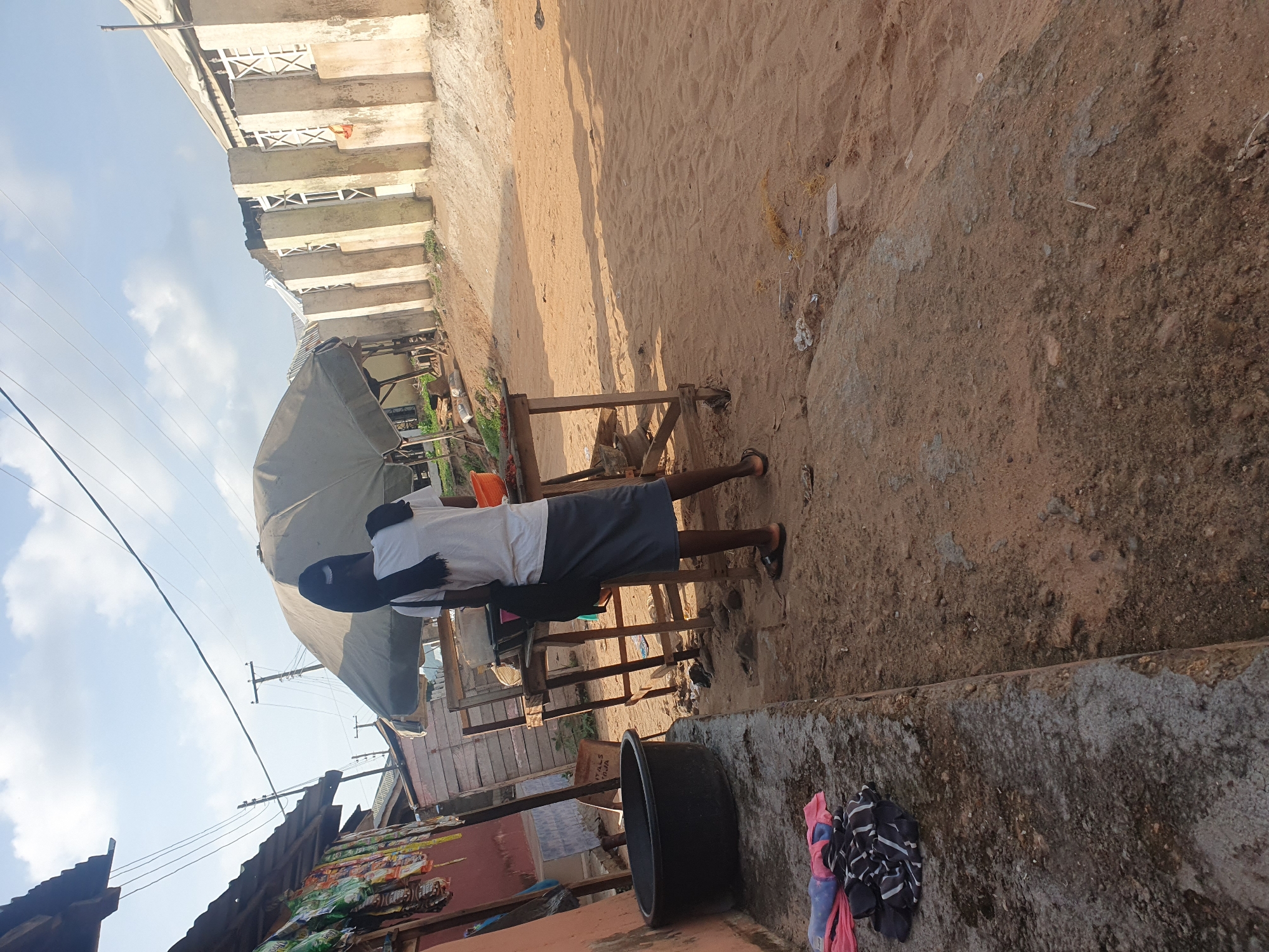 A person wearing a black headscarf, white t-shirt, and gray skirt stands at a wooden market stall under a large umbrella in Lokoja, Nigeria. The stall has a wooden frame and tabletop. A large black basin is to the left of the stall, and a pile of folded clothes is on the concrete edge in front of it. Behind the stall, a building with white-framed windows and concrete pillars is visible. The ground is sandy, with some debris scattered around. Overhead, power lines stretch across a cloudy sky.