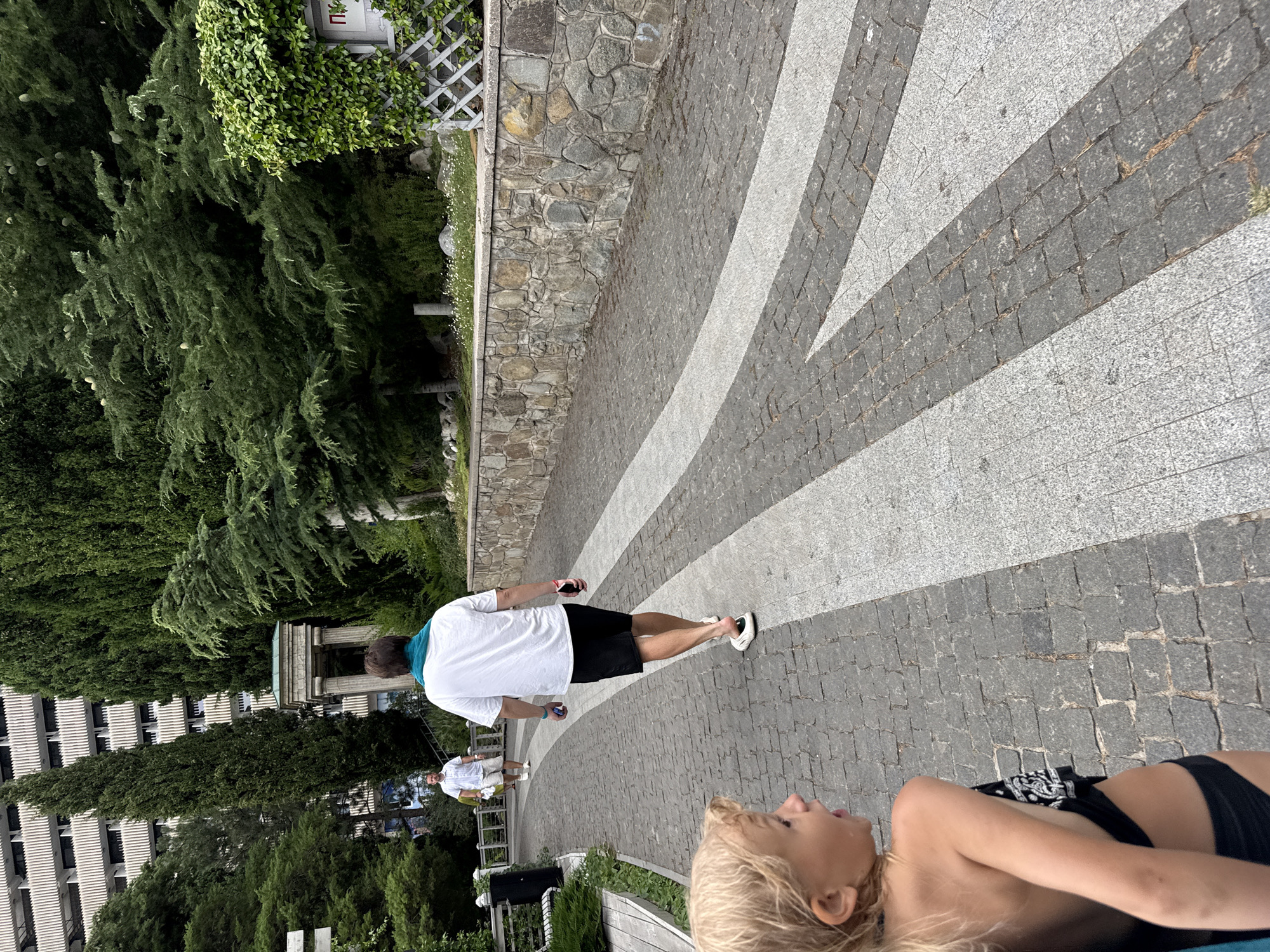 The image depicts a sunny day in Yalta, Ukraine. A man in a white t-shirt and black shorts walks away from the camera on a paved pathway. He is wearing a turquoise scarf around his neck and white sneakers. Another man, wearing a white shirt and shorts, walks in the distance on the same path. In the foreground, a person with blonde hair lies down, looking up.
The pathway is made of grey and white cobblestones, arranged in a pattern that suggests a walkway or plaza. Lush green trees and dense foliage line the left side of the pathway, with a stone wall and a white lattice structure visible behind them. In the background, a large, modern building with white horizontal louvers rises above the trees. The overall atmosphere appears relaxed and casual, consistent with a warm, outdoor setting. The lighting suggests it is daytime, likely mid-morning or afternoon, given the bright sunlight and shadows. There is no visible text in the image.