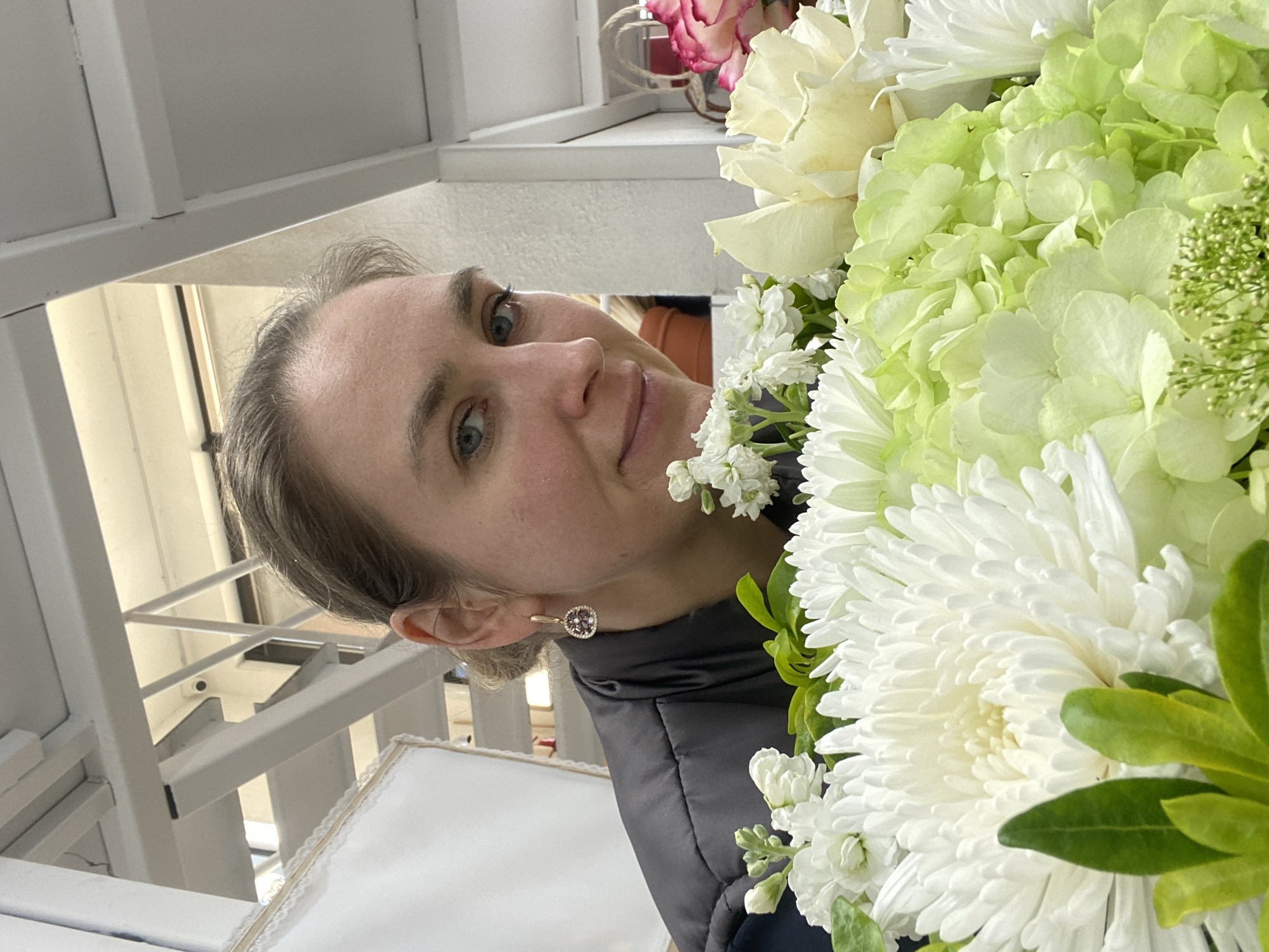 A fair-skinned woman with graying hair and blue eyes poses behind a large bouquet of white and light green flowers. She is wearing a dark jacket and gold earrings. The background features a white railing and glimpses of a building interior. The presence of the flowers suggests a possible location related to floristry or an event decorated with floral arrangements. The location of Yekaterinburg, Russia is not discernible from the image.