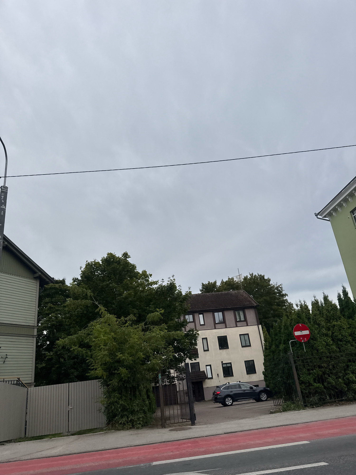 The image depicts a residential street in Tallinn, Estonia, under an overcast sky. The foreground shows a street with a red-marked lane, likely a bike lane or a pedestrian crossing. To the left, a two-story building with light green siding and white horizontal trim is visible, partially obscured by a grey corrugated metal fence. A lamppost extends from the upper left corner.
In the middle ground, a multi-story building with a light beige facade and darker brown trim around the windows and roofline stands. The building has multiple windows and appears to be an apartment complex. A dark-colored SUV is parked in front of this building. Behind and to the right of this building, a dense line of dark green evergreen trees forms a natural barrier. To the far right, another building, painted a bright green with white decorative molding near the roof, is partially visible.
A red and white circular traffic sign, indicating a prohibition, is mounted on a metal pole to the right of the evergreen trees. The overall environment suggests a suburban or residential area within Tallinn. There are no people visible in the frame.