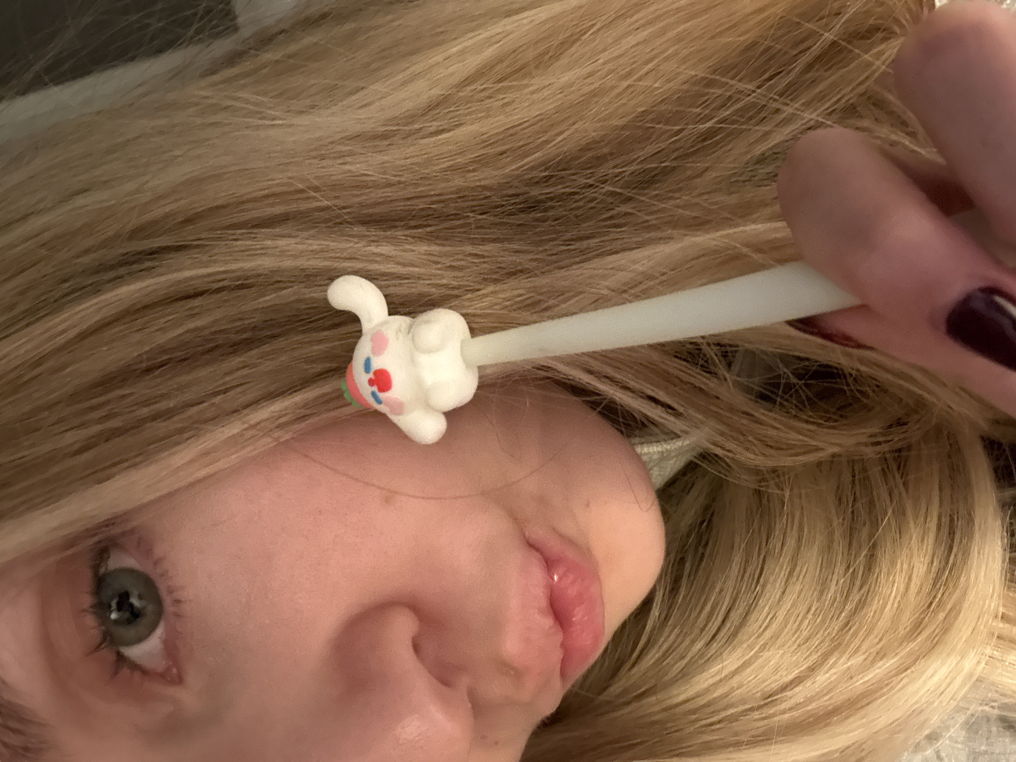 A close-up, tilted portrait of a person with light blonde hair is shown. The person's face, including their left eye, nose, and mouth, is visible. The person's right hand and fingers holding a white, cylindrical object with a small, white, bunny-shaped decoration on the end are also visible. The background appears to be blurry and neutral in tone. No information suggests the location is in Mexico City, Mexico.
