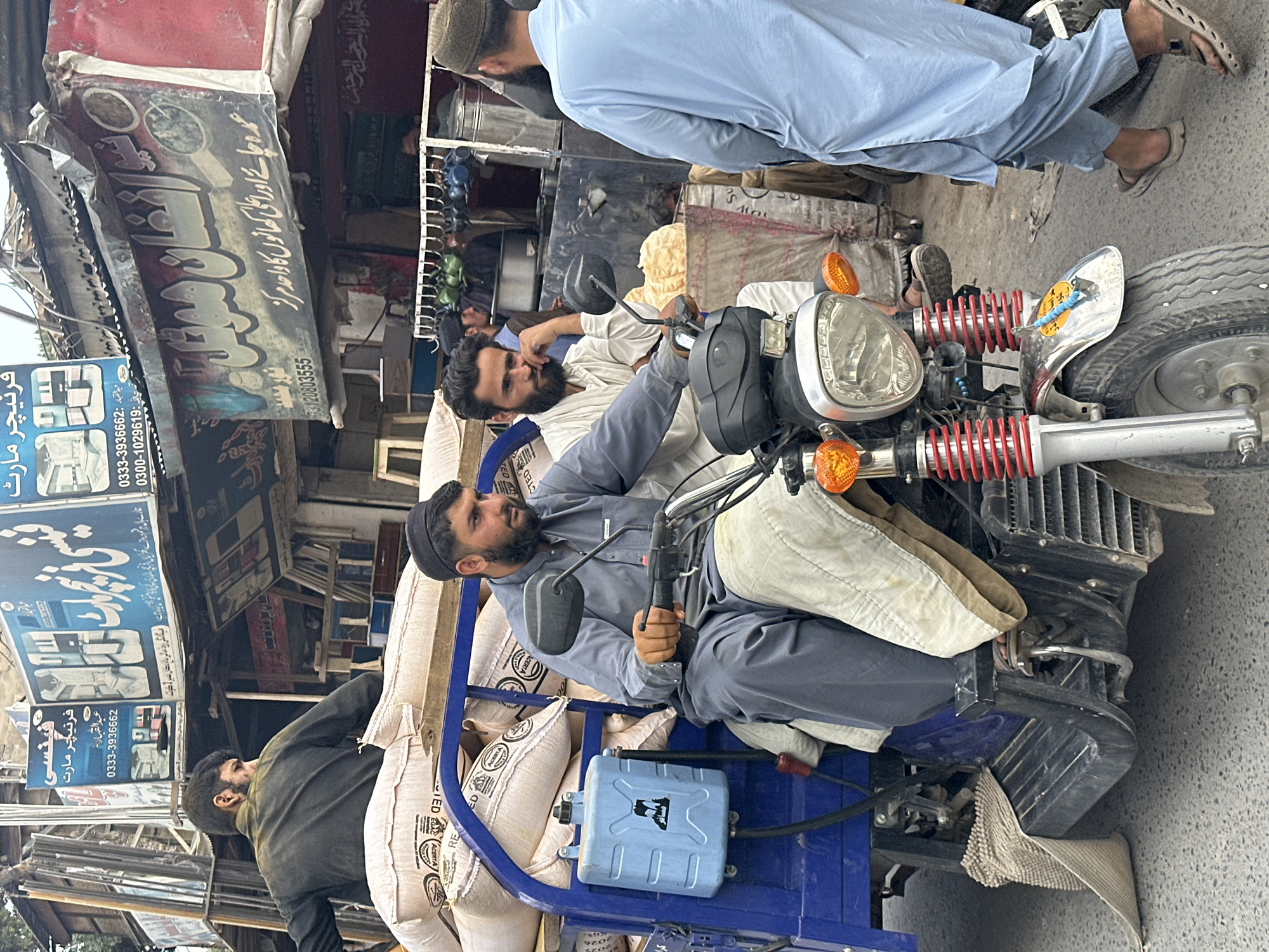 A bustling street scene in Quetta City Tehsil, Pakistan, showcases a three-wheeled motorcycle loaded with sacks. The driver, a middle-aged man with a beard and wearing a traditional cap, is sitting on the vehicle, ready to go. Another man, also bearded, is sitting behind him on top of the sacks, seemingly waiting to be transported. A third man in a light blue outfit walks by. The street is surrounded by shops with cluttered storefronts and advertisements, including signage for furniture and tea shops. The environment has the characteristics of a busy commercial area.