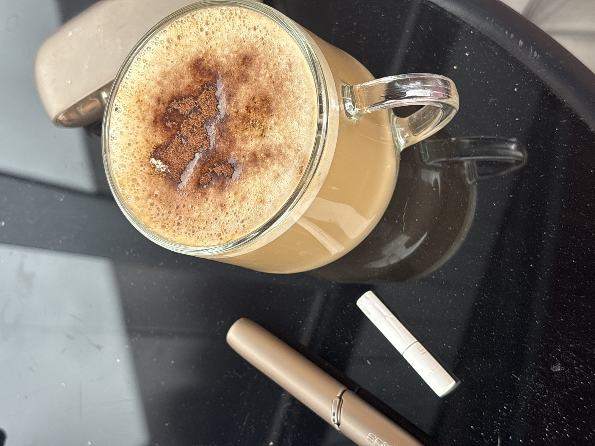 A top-down, medium shot shows a glass mug of frothy coffee with a sprinkle of cocoa powder on top, positioned on a dark, reflective surface, likely a table. To the left of the mug, there is a golden electronic smoking device and a white cigarette stick with an orange tip. In the background, a portion of a similar electronic device is visible. The scene appears to be indoors, likely in a domestic or public setting within the city of Semiluki, Russia. The lighting suggests it is daytime.