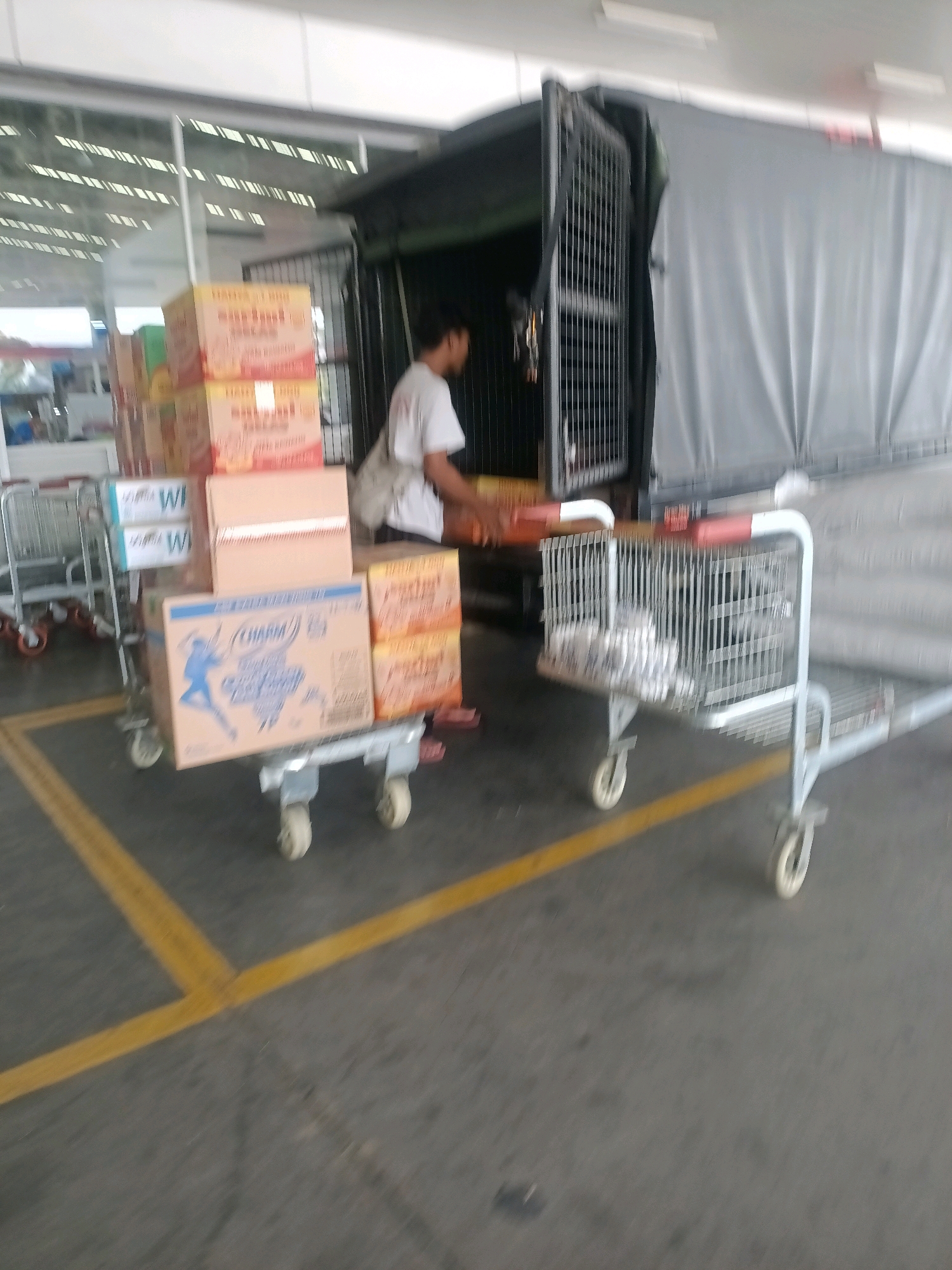 In Jaten, Indonesia, a man in a white t-shirt is seen partially within the rear opening of a dark-colored truck. The truck has a grey tarp covering its load and a metal cage structure visible at the opening. In the foreground, several shopping carts are present. One cart holds stacked boxes, including boxes with blue and yellow labels that read "WH" and another box featuring a blue silhouette of a person with the text "CHARM". Another cart in the mid-ground contains what appear to be wrapped packages, possibly paper towels or toilet paper. The ground is paved with a grey surface and marked with yellow lines, indicative of a loading dock or parking area. The environment appears to be an outdoor commercial or warehouse setting with a building visible in the background.