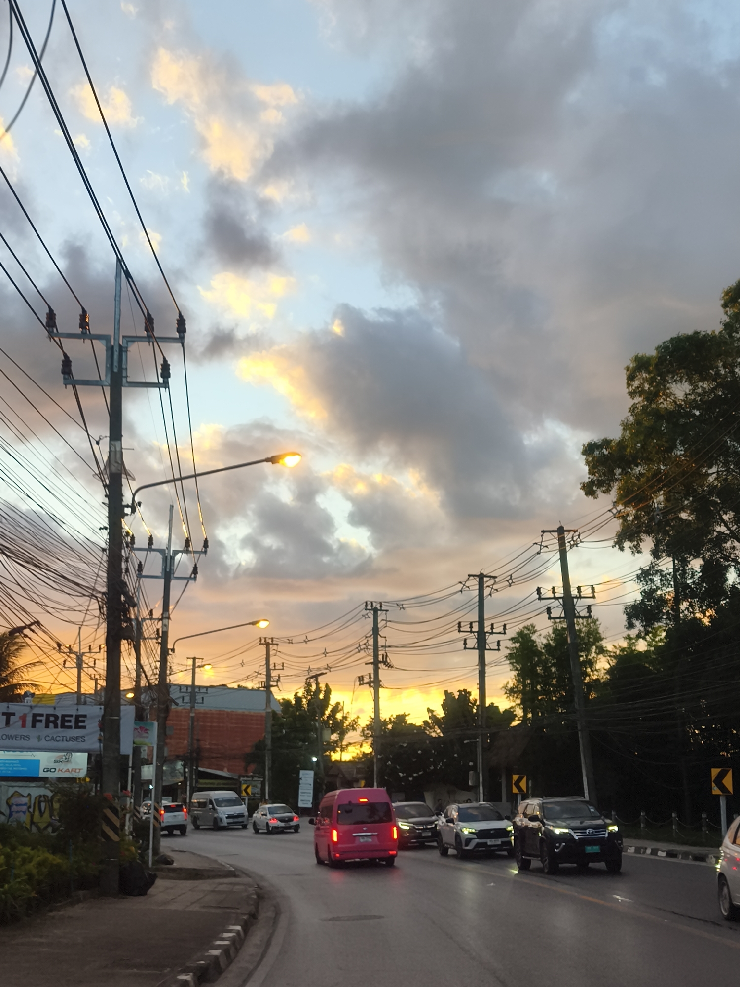 This image captures a street scene in Baan Bo Phut, Thailand, during what appears to be sunset. The sky is filled with dramatic clouds, tinged with hues of orange, yellow, and blue, suggesting the end of the day. Power lines crisscross the frame, supported by numerous utility poles, a common sight in many Southeast Asian locales.
The road is moderately busy with traffic. Several cars, including a white van, a dark SUV, and a bright pink van, are visible. A white car is seen in the far right, possibly moving away from the viewer. Streetlights are illuminated, casting a warm glow on the road and adding to the evening atmosphere. Trees line the right side of the road, their leaves dark against the colorful sky.
On the left side of the road, there's a sidewalk with some vegetation and a sign that reads "TI FREE" and "FLOWERS CACTUSES". Below this, a smaller sign says "GO KART". The presence of these signs, along with the general streetscape, points to a commercial or semi-urban area. The time of day is clearly indicated by the sunset sky and the activated streetlights. The weather appears to be partly cloudy, with patches of clear sky visible. There are no people prominently featured in the image, making the focus on the road, the vehicles, and the atmospheric sky.