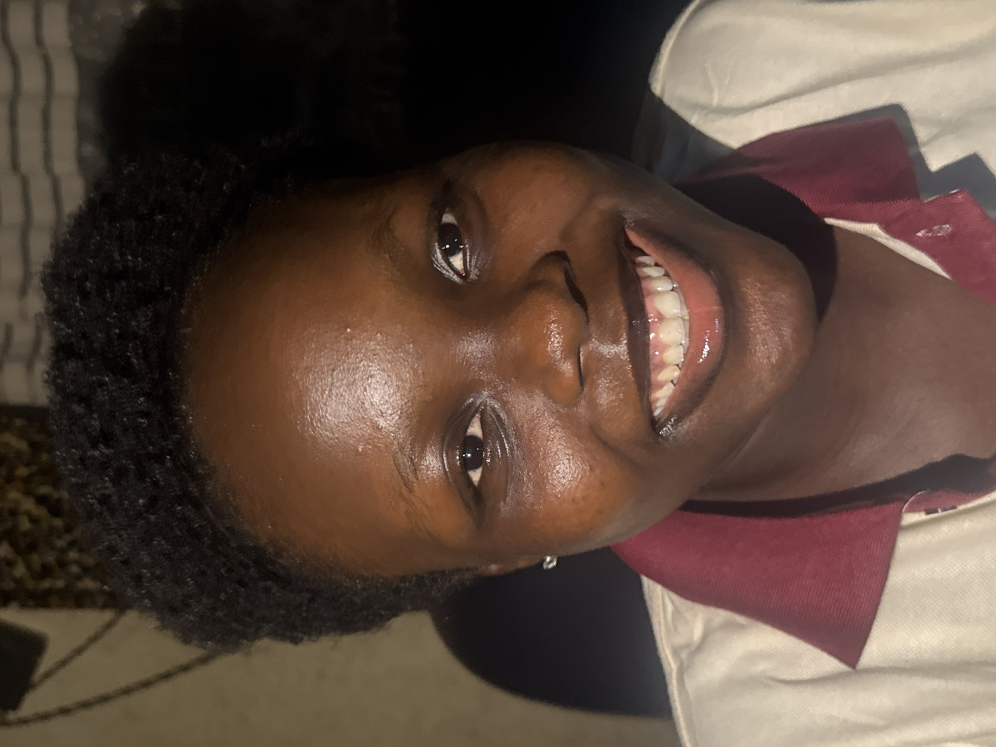 A close-up, dimly lit, rotated portrait shows a young Black woman with short, dark, natural hair, smiling broadly. She is wearing a collared shirt, with the collar being a burgundy color and the rest of the shirt a light color. The background appears dark and indistinct. The image does not provide sufficient information to verify the location of Onitsha, Nigeria.