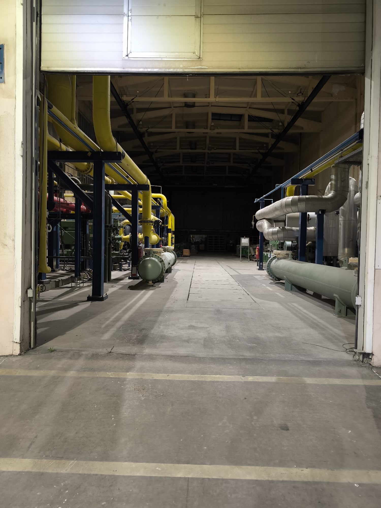 A vertical shot captures the interior of a dimly lit industrial building, possibly a factory or warehouse, viewed from an open doorway. The foreground is dominated by a concrete floor marked with yellow lines. To the left, a complex network of industrial pipes, predominantly bright yellow and supported by dark blue structures, runs along the wall. Some red pipes are also visible. A green cylindrical tank is situated near the pipes. The right side of the image features larger, silver-colored pipes with insulation, and another long, olive-green cylindrical tank rests on supports. The background recedes into darkness, with the ceiling structure and some lighting elements visible. The environment appears to be a functional industrial space, free of people, with a focus on machinery and infrastructure.