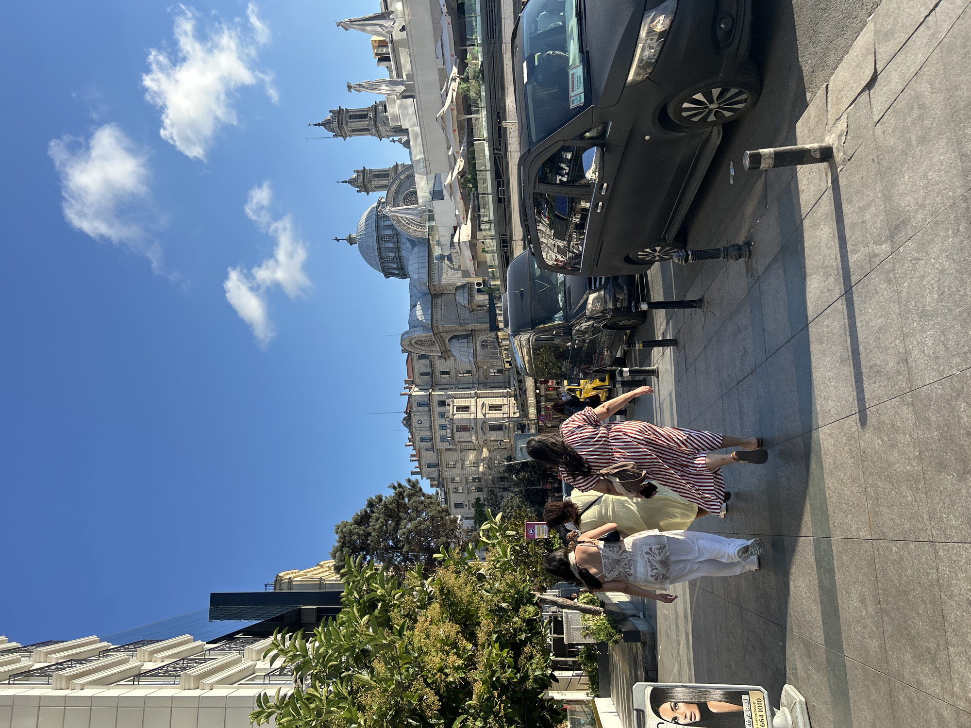 In Beyoğlu, Turkey, on a sunny day, three women walk on a paved sidewalk. The woman in the foreground wears a red and white striped dress and carries a brown bag. To her left, another woman in a light yellow dress walks alongside. The third woman, on the far left, is dressed in white and wears white sneakers. Behind them, a black van is parked on the street, and further down, a yellow taxi is visible. The street is lined with trees and buildings, including a grand structure with domes and spires in the background. A billboard featuring a woman's face is placed on the left side of the sidewalk. The sky is a clear blue with scattered white clouds.