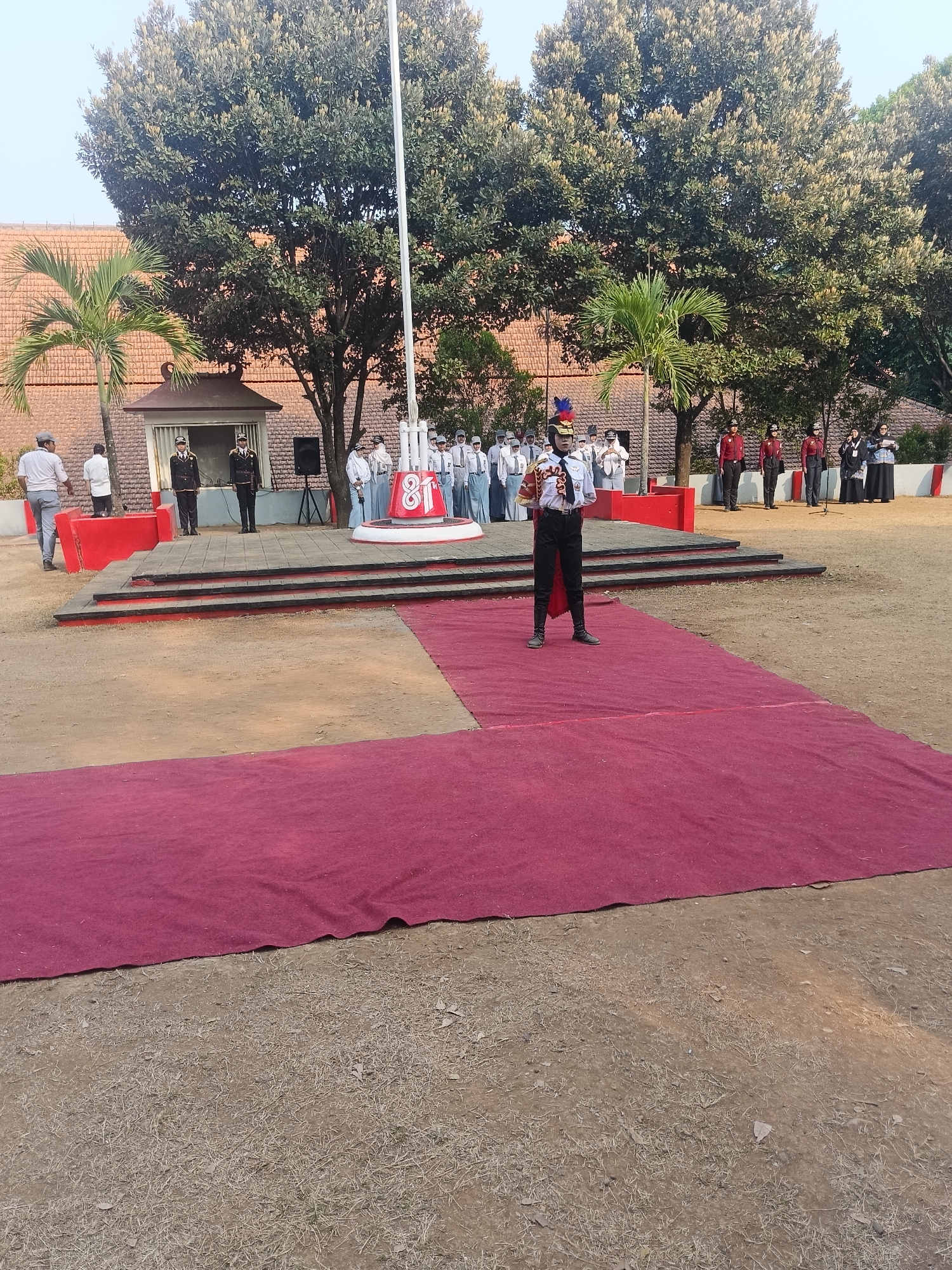 In Pengkol, Indonesia, a group of students are gathered for what appears to be an outdoor ceremony or assembly. The scene is set on a dirt ground covered by a large red carpet in the foreground. Behind the carpeted area, a raised platform is visible with several steps leading up to it. On this platform, a contingent of individuals in uniform are standing, including two in dark uniforms with hats and epaulets, and a group of students in white shirts and grey pants or skirts, some with headscarves. To the right of this group, another line of individuals in red shirts and dark pants are present. In the center of the platform, a flagpole stands tall. A person in a ceremonial uniform with a plumed hat is standing on the red carpet in the foreground, facing the group on the platform, and appears to be addressing them or preparing to do so. The background features mature trees with dense foliage and a building with a tiled roof, suggesting an institutional setting like a school or community center. The overall atmosphere suggests a formal event taking place during daylight hours.