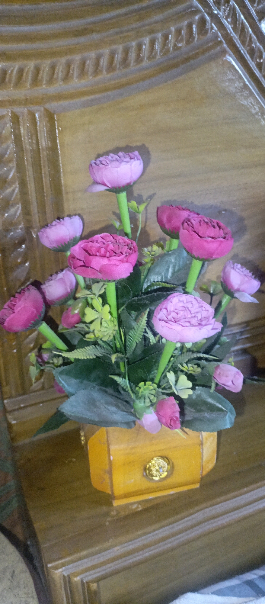 A close-up, vertical, eye-level shot captures a faux flower arrangement featuring multiple pink and purple roses in various stages of bloom, set against a backdrop of rich, textured wooden furniture. The flowers, with their fabric petals, are arranged in a small, bright yellow wooden pot adorned with a circular, ornate gold accent in the center. The wooden furniture exhibits a polished, aged appearance with carved details and horizontal lines, suggesting a traditional or antique style. The lighting is soft, casting gentle shadows that highlight the textures of both the flowers and the wood. The scene appears to be indoors, possibly in a home or establishment in Gazipur, Bangladesh, as indicated by the context of previous interactions, though there are no identifiable landmarks or specific indicators of this location within the image itself. The composition focuses solely on the floral arrangement and the immediate wooden surface it rests upon, creating an intimate still life.