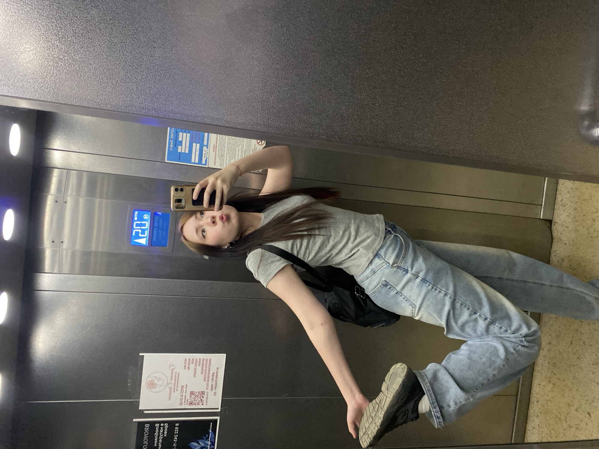 A young woman is taking a selfie in an elevator. She is wearing a gray t-shirt, light blue jeans, and black sneakers. She has a black bag slung over her shoulder. The elevator has an electronic display showing "20" and advertisements/notices attached to the wall. The advertisements contain Russian text. The elevator's interior is primarily gray metal. The location appears to be a building in an urban setting. The city of Unknown, Unknown cannot be determined from the image.