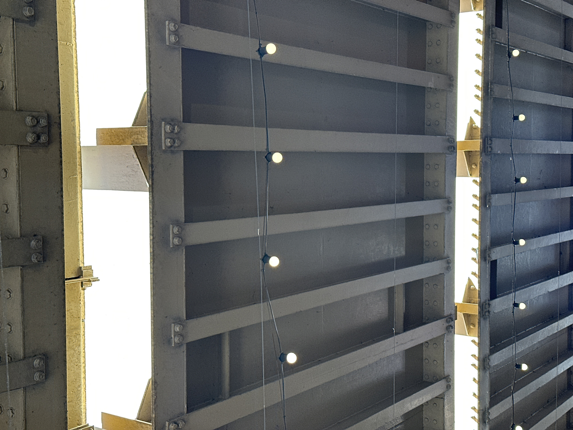 The image depicts a close-up, upward view of the underside of Tower Bridge in London, United Kingdom. The bridge's metal framework is visible, with bolts and crossbeams connecting the structural elements. There are rows of small, spherical lights suspended by thin wires. Gaps in the structure allow bright light to filter through, suggesting daylight. The metalwork appears to be painted in a neutral color.