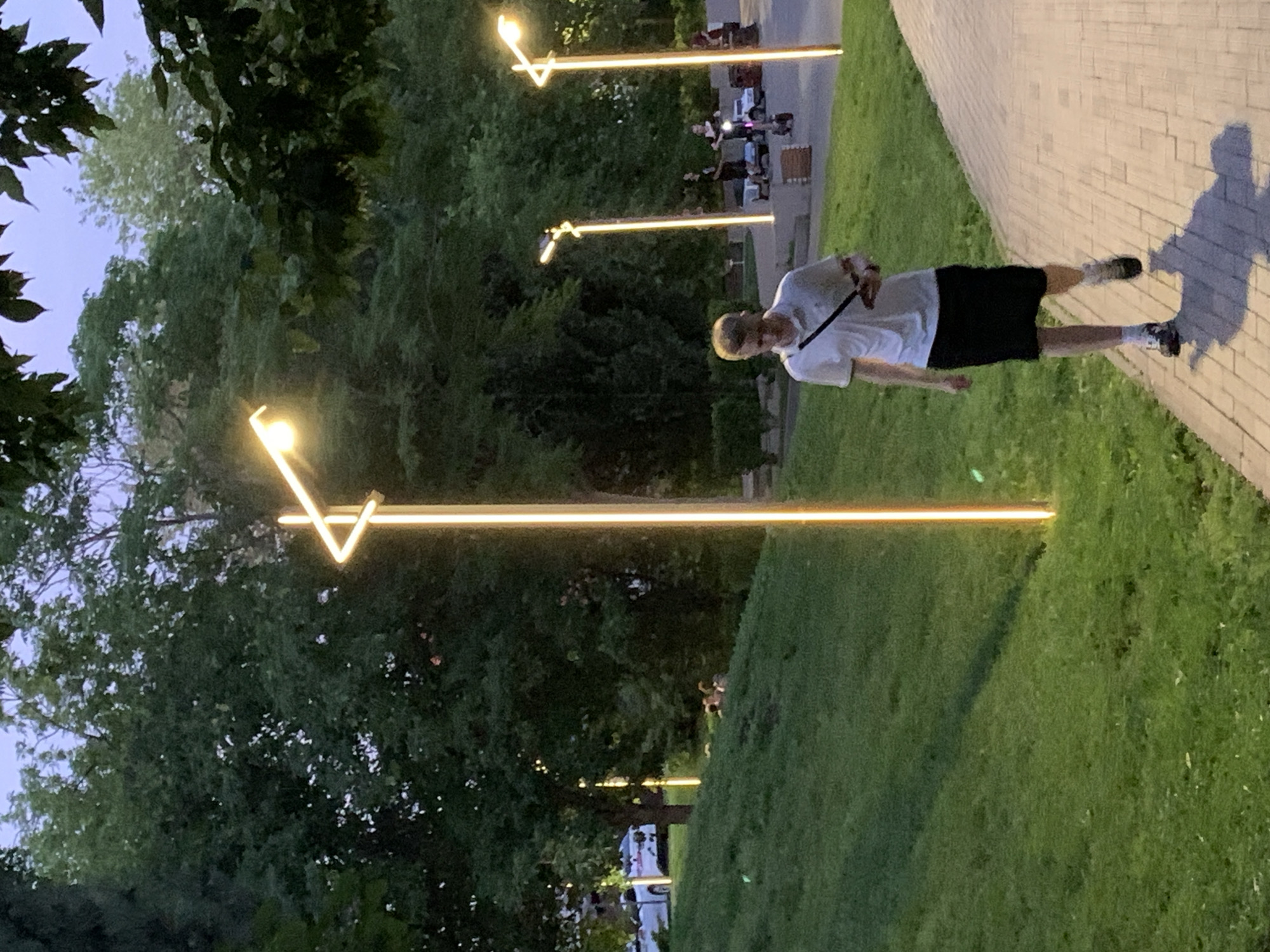 A young man with blond hair, wearing a white polo shirt and a black skirt, walks on a paved path next to a grassy hill. He is holding a phone in his right hand. Modern streetlights with glowing orange lines illuminate the path and the surrounding trees. In the background, several people are visible, some of whom appear to be riding electric scooters and other personal mobility devices. A white car is parked on the street in the distance, beyond the grassy hill. The scene is set in a park-like environment with lush green grass and dense trees, suggesting an urban park or recreational area. The sky is a muted twilight blue.