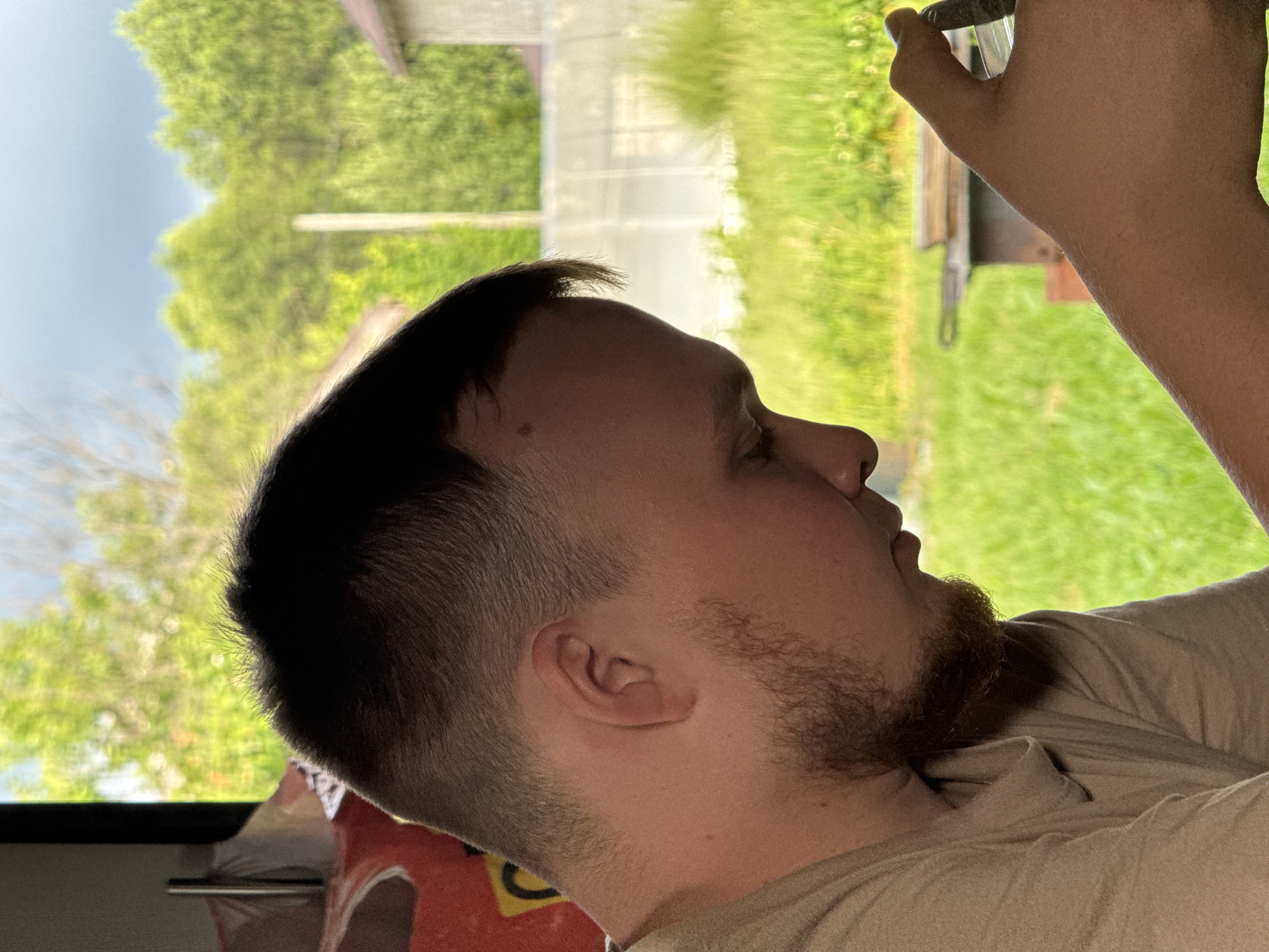 The image depicts a male individual in profile, facing right and looking downwards, against an outdoor background in the city of Krasnokamsk, Russia. The man has short, dark hair with a faded cut on the sides, and a reddish-brown beard. He is wearing a light brown or tan short-sleeved shirt. A small, dark mole is visible on his forehead. His right hand is partially visible in the lower right corner, holding a small, clear, cylindrical object. The background, viewed through an opening, consists of vibrant green grass and trees under a light blue, possibly cloudy, sky. Distant structures include a light-colored building with a dark roof on the right, and a white, possibly netted or plastic, structure resembling a greenhouse or fencing further back. A wooden utility pole stands in the middle distance. A reddish object with yellow accents is partially visible on the left side, behind the man's head, suggesting an indoor setting for the foreground subject.
