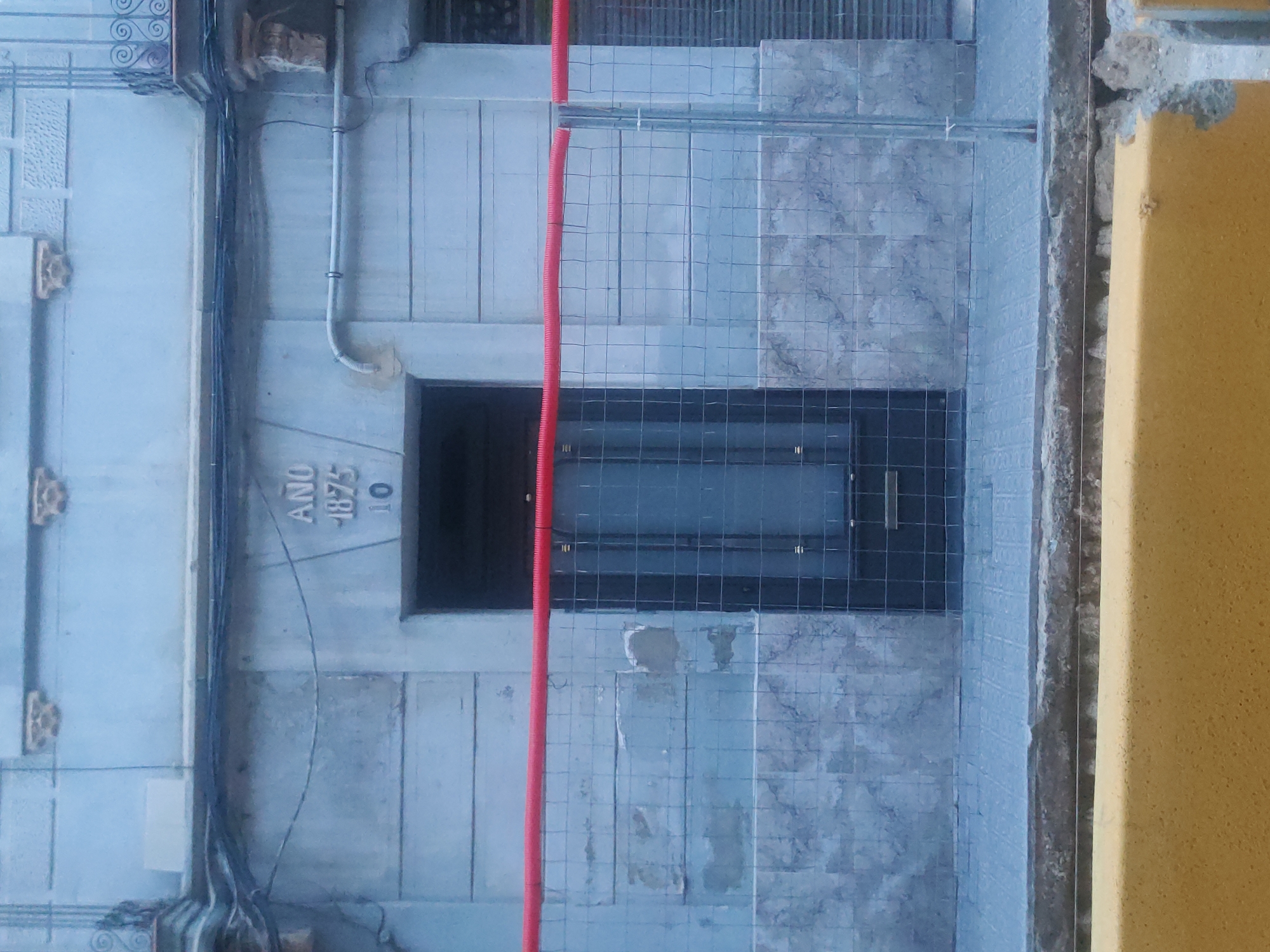 The image shows the facade of a building. Above a dark doorway, there is an inscription that reads "AÑO 1875 10". The building's exterior is made of light-colored stone blocks. A metal fence with a red pipe running along the top is in front of the doorway. The ground in front of the fence is paved with square tiles. A yellow barrier is visible in the foreground. The scene appears to be in an outdoor urban environment.