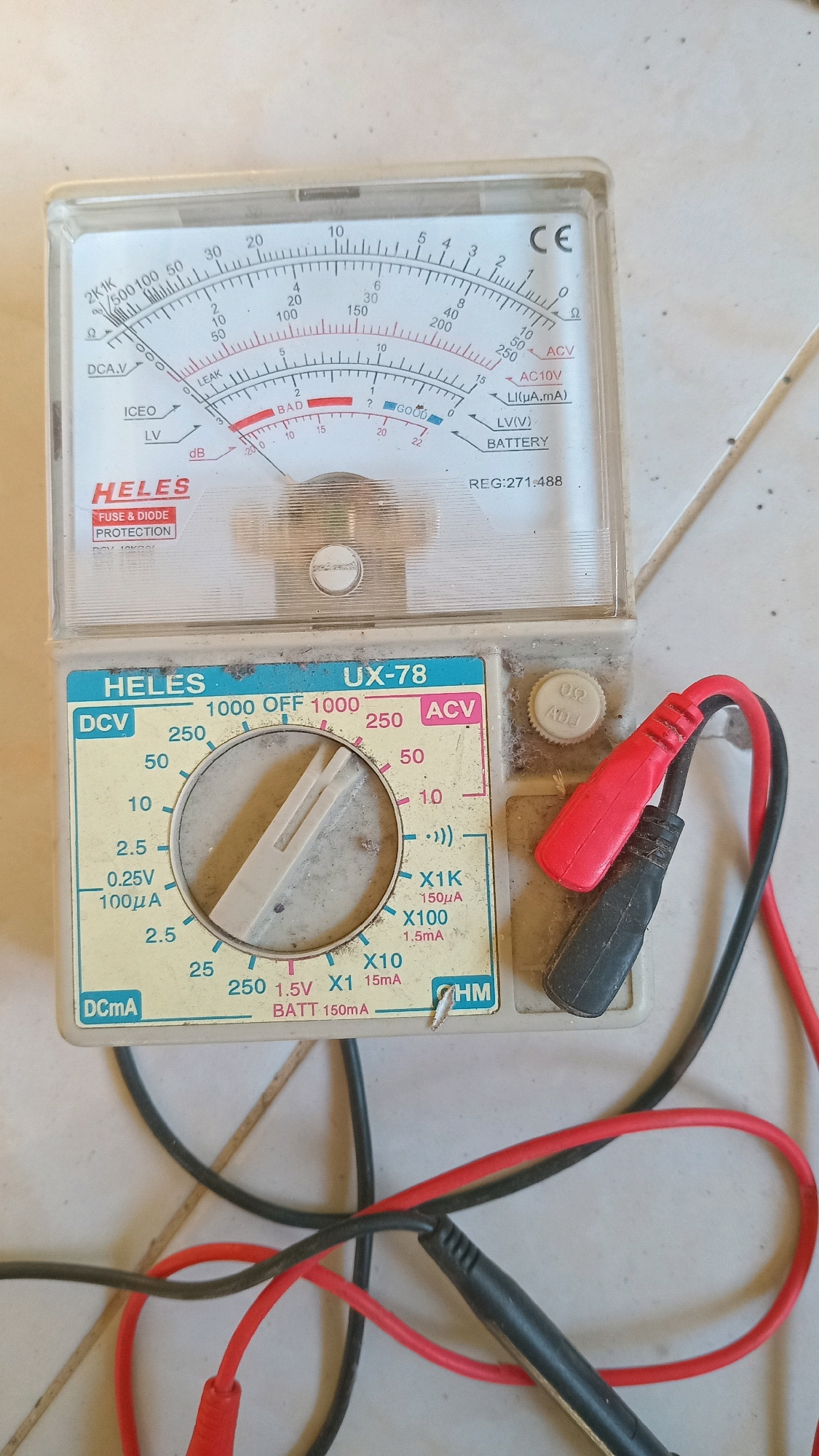 An analog multimeter, model UX-78 by HELFS, is pictured lying on a tiled floor in Randuagung, Indonesia. The device has a white casing with a clear plastic display cover. The dial is set to the "1000 OFF 1000" position, indicating it is not currently measuring anything. Red and black test leads are connected to the multimeter, with the red lead positioned over the ACV setting and the black lead over the DCV setting. A white adjustment knob labeled "Ω ADJ" is visible to the right of the dial. The multimeter displays various scales for measuring voltage, current, resistance, and decibels. The tiled floor appears to be weathered and has some debris scattered across it.
