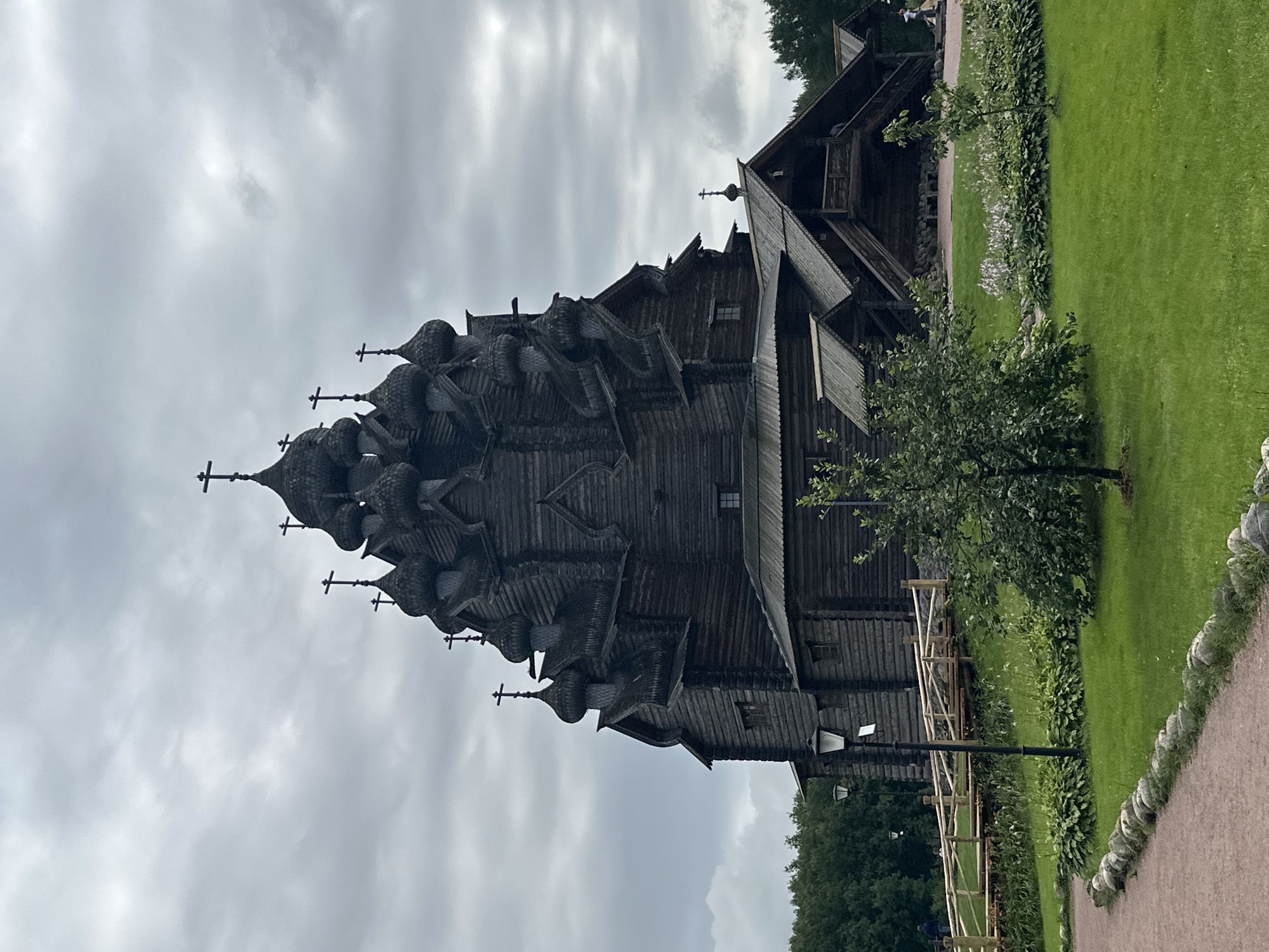 The image shows a large, dark wooden church with multiple onion domes and crosses against a cloudy sky. The church is situated in a grassy park area with trees and landscaping. A wooden walkway with railings leads up to a covered entrance on the right side of the church. A lamppost stands in the foreground to the left of a pathway. The church is located in Nevskii Parkleskhoz, Russia. There are no people clearly visible in the immediate vicinity of the church, though a few figures can be seen in the distance on the walkway. The overall atmosphere is somber due to the overcast weather and the dark color of the wooden structure.