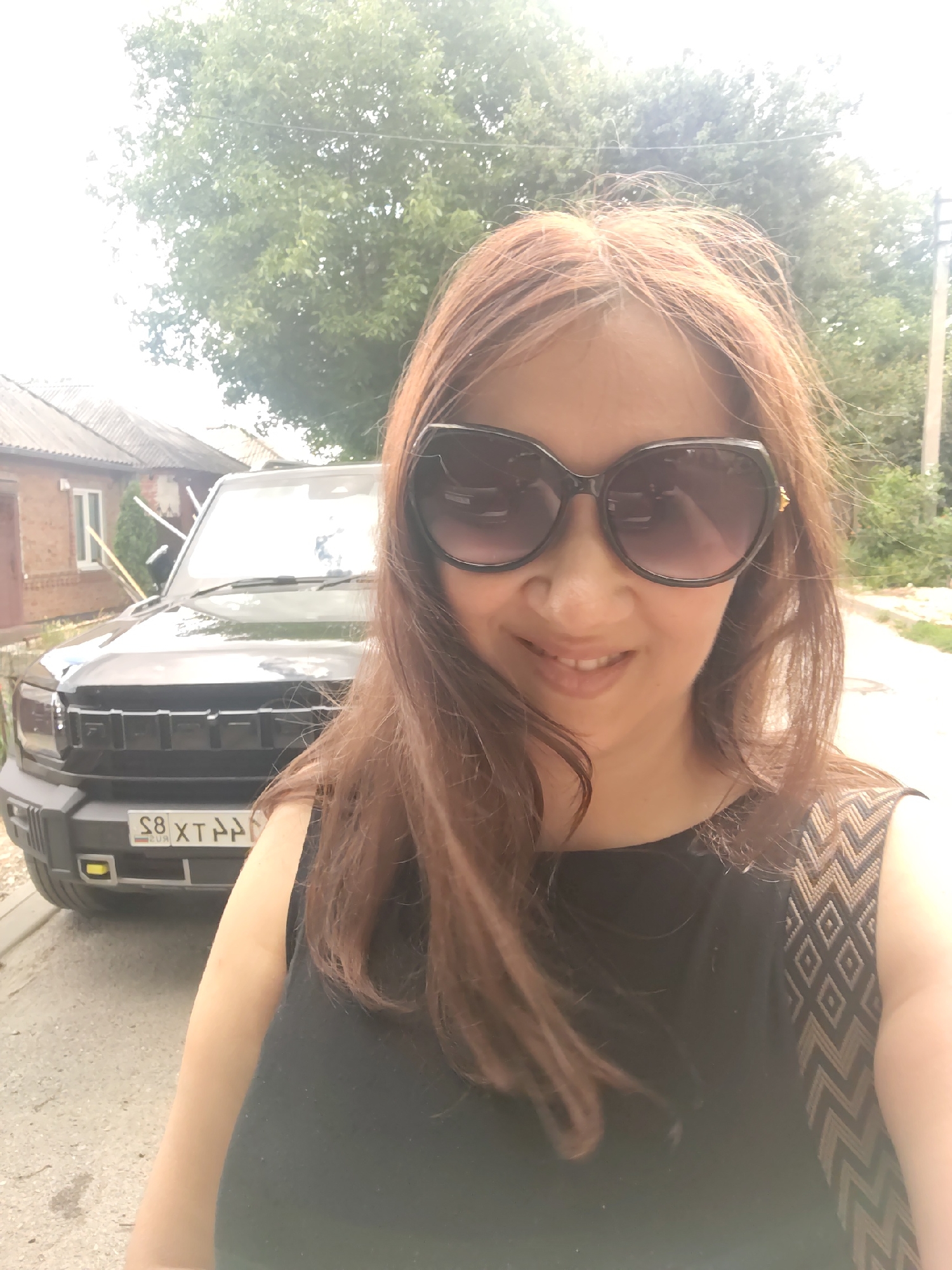 The image is a selfie of a woman wearing sunglasses and a black top with a patterned detail on the shoulder. She has long, reddish-brown hair. Behind her, a black SUV is parked. The license plate of the SUV is visible and reads "S8 XT AA". The background shows a building with a grey tiled roof, trees, and a street. The woman is smiling. The scene appears to be outdoors in a residential area, possibly a driveway or street. Based on the license plate format, the location is likely Russia. The structure of the license plate and the characters are consistent with Russian license plates. Therefore, this image may have been taken in Rostov-on-Don, Russia.