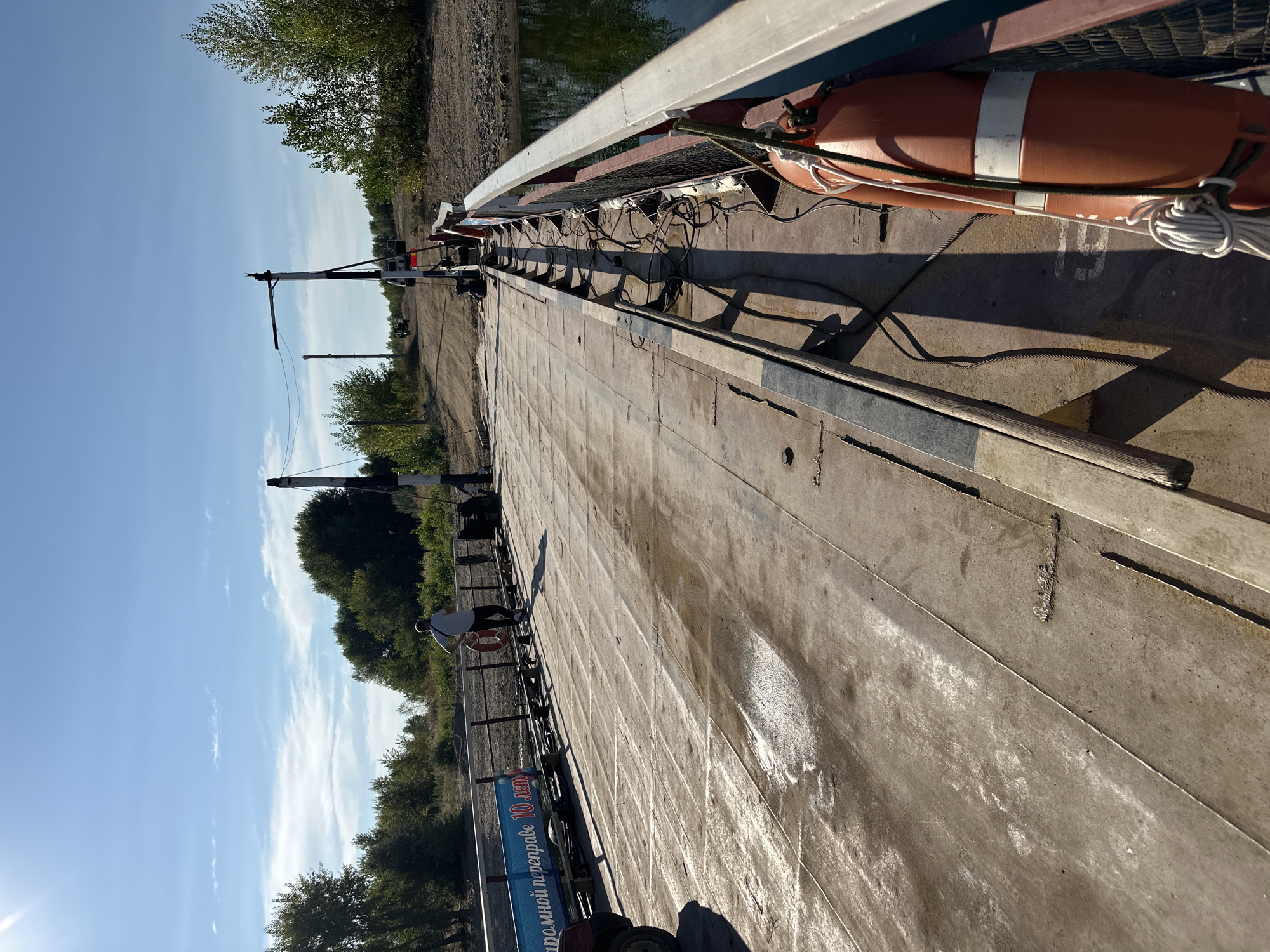 The image shows a ferry platform on a body of water, likely a river, on a sunny day. The platform is made of weathered metal plates and appears to be carrying vehicles, with at least one vehicle partially visible on the left. A person is standing near the railing in the middle of the platform, facing away from the camera, holding what appears to be a phone or a small device. Another person or a figure in a light-colored shirt is visible further down the platform. There are safety buoys attached to the railing. The surrounding environment consists of trees and vegetation along the bank, with a dirt road leading away from the ferry on the far bank. The sky is blue with scattered clouds. The scene is likely located in or near Primorsk, Russia, given the context provided.