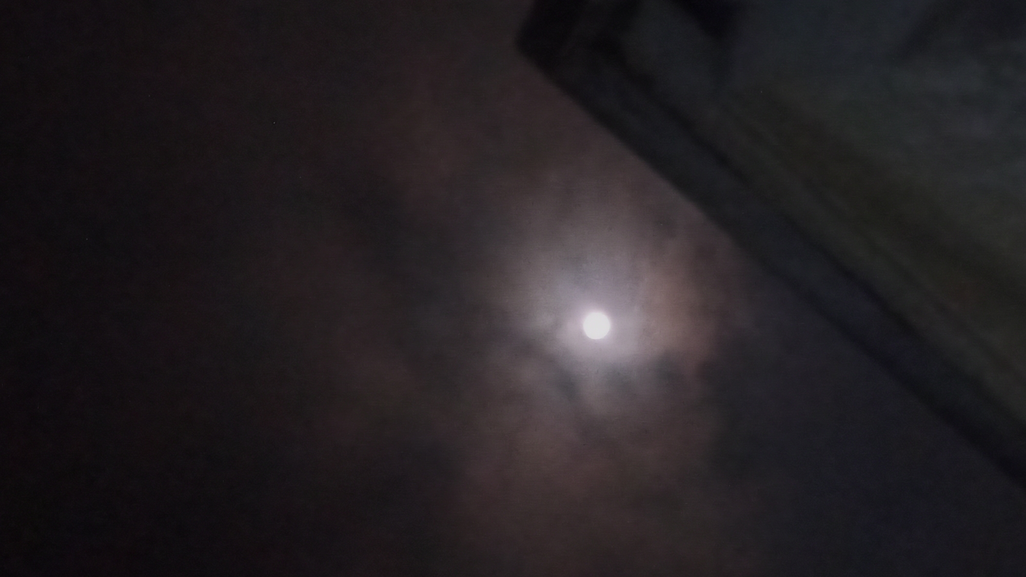The image captures a nighttime scene in Tuba, Indonesia, focusing on a hazy full moon partially obscured by clouds. In the lower right portion of the frame, the edge of a building's roofline is visible, rendered in dark tones. The overall atmosphere is dim and somewhat mysterious due to the low light conditions and the cloudy sky. There are no people or discernible actions taking place in the image.