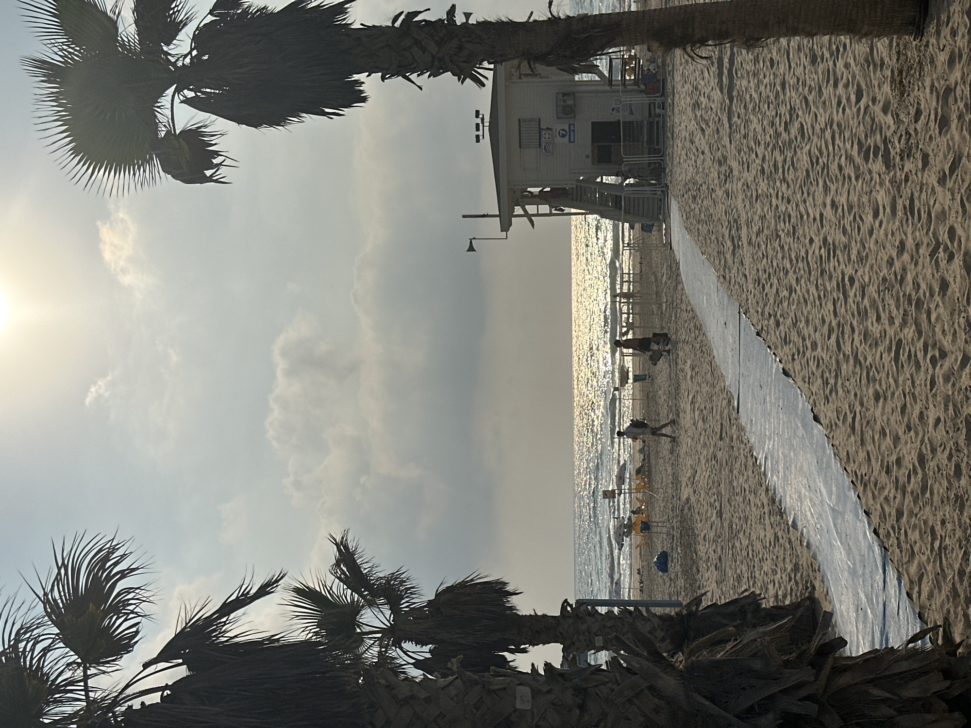 This is a daytime, eye-level, vertical shot of a beach in Bat Yam, Israel, featuring palm trees framing the scene. In the foreground, a makeshift walkway of white plastic sheeting is laid over the sand, leading towards the ocean. To the right, a white lifeguard station stands with a set of stairs. Silhouettes of people are visible on the beach, some walking and others near beach chairs and umbrellas in the mid-ground. The sea is calm with a shimmering reflection of the sun, and the sky is partly cloudy. The overall lighting suggests it is late afternoon.