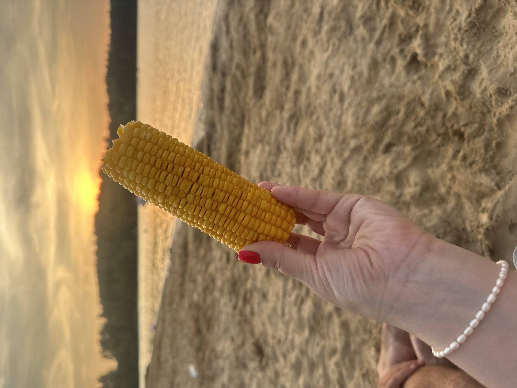 A person's hand, with red nail polish and a pearl bracelet, holds a cob of corn against a blurred background of a beach and a body of water at sunset. The sand is light brown and textured. The water is calm and reflects the orange and yellow hues of the sky. Trees form a dark silhouette along the far bank of the water. The sky is filled with wispy clouds tinged with sunset colors. The location is the Yauza River in Lytkarino, Russia.