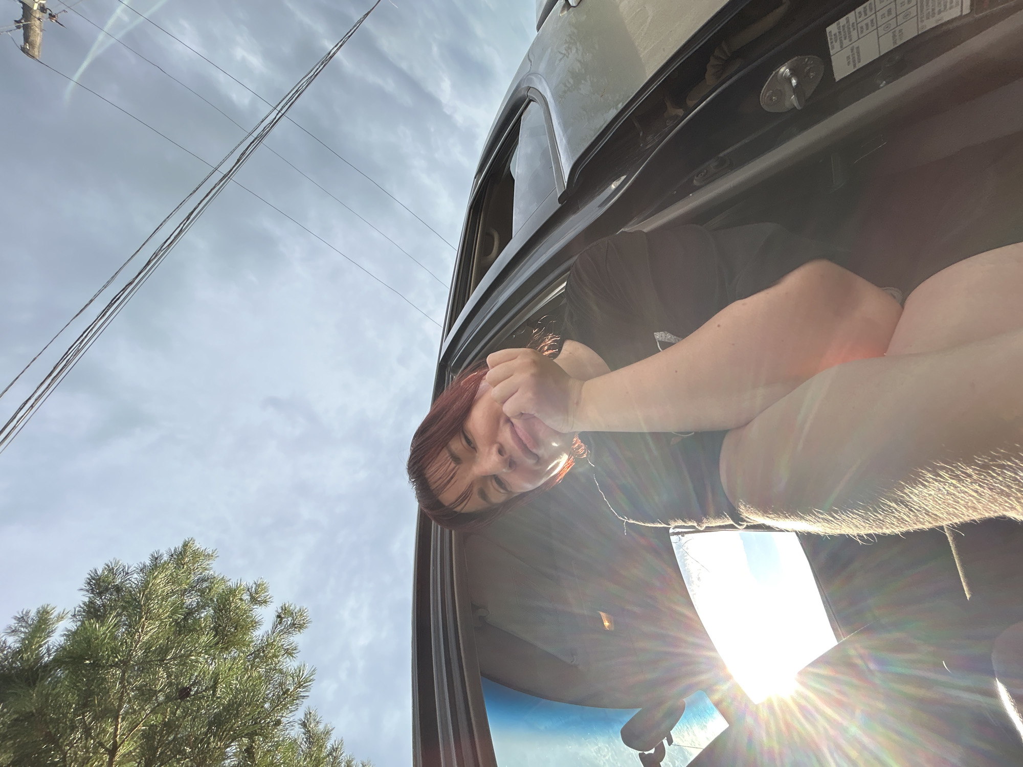 In a scene outdoors, a young person with short, dark red hair and bangs is sitting inside the open door of a vehicle. They are leaning their chin on their hands, with their arms bent. The person is wearing a dark t-shirt. The sun is shining brightly, creating a lens flare that extends from the lower left of the frame across the image. In the background, a tree with green needles is visible in the upper left corner, and power lines stretch across the sky, which is partly cloudy. The ground is not visible. The image was taken in the city of Foky, Russia.