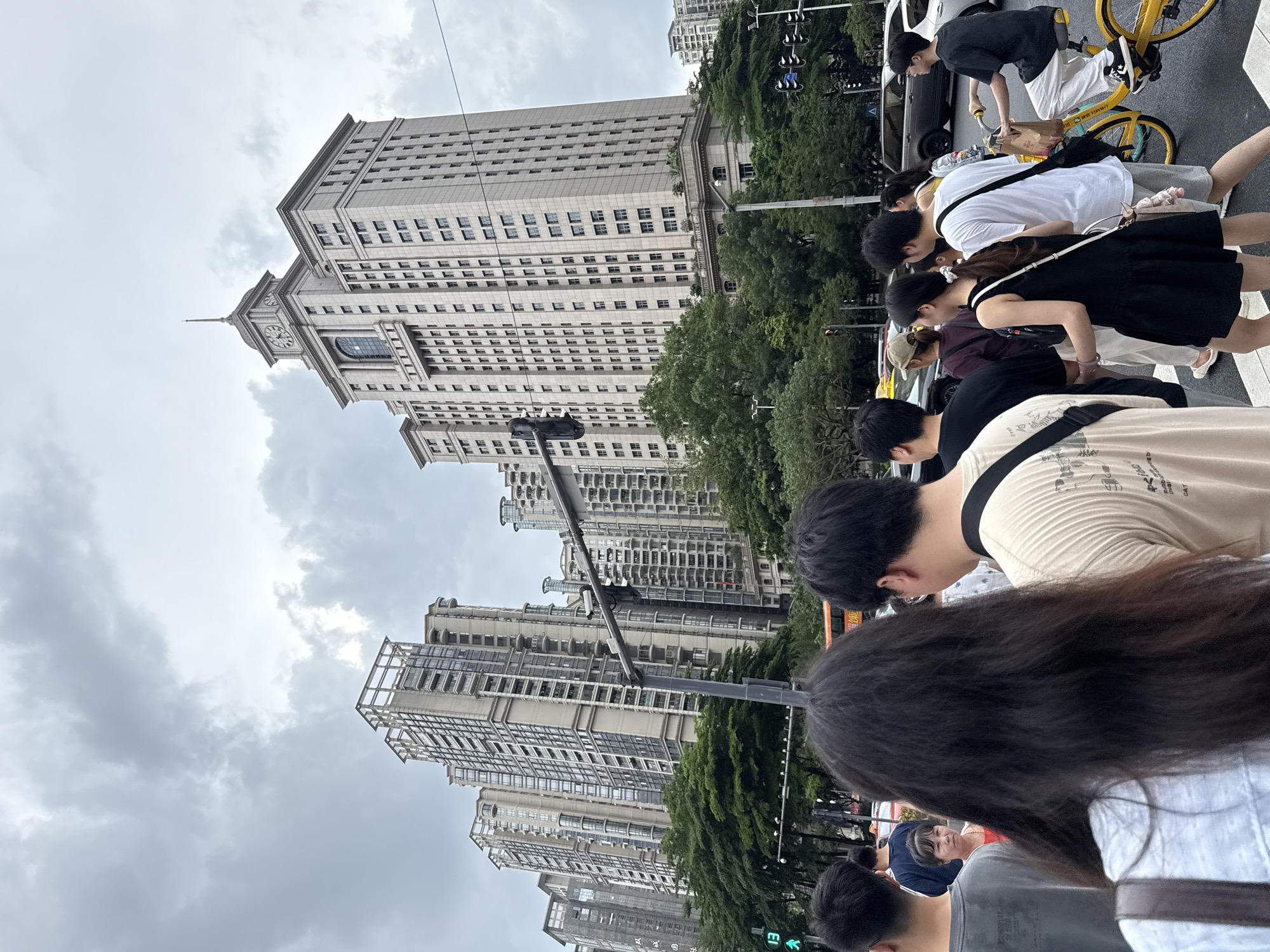A group of people are walking on a street with tall buildings in the background. The buildings are modern with many windows. Trees line the street. A yellow bicycle is visible. Some people are looking at their phones, while others are walking with purpose. The sky is cloudy, suggesting it might be an overcast day. The people are dressed in casual clothing. There is no text clearly visible in the image. The overall scene depicts an urban environment in China, possibly during the daytime, with people engaged in everyday activities.