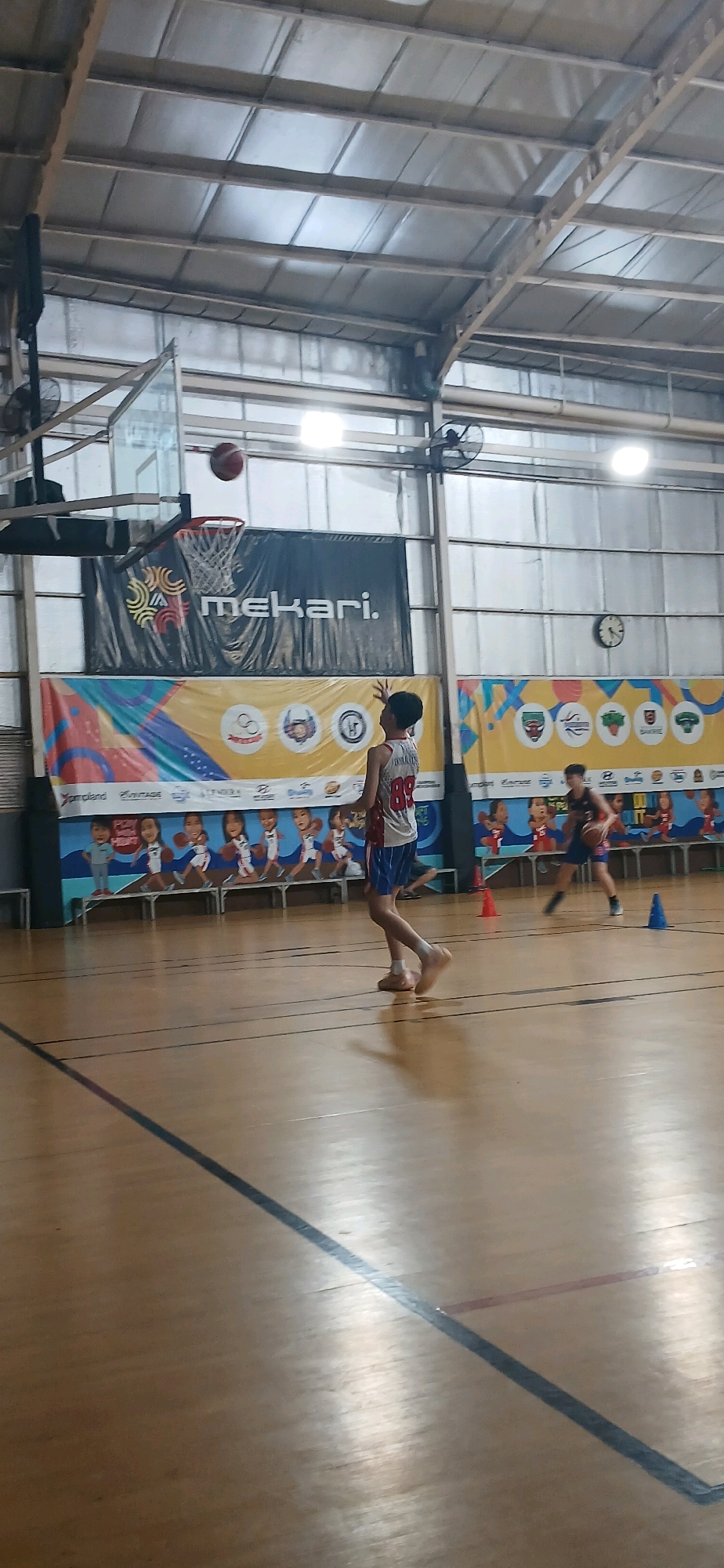 In an indoor basketball court in Cirebon, Indonesia, two young male basketball players are engaged in a game. The court floor is made of polished wood. In the foreground, one player, wearing a white and red jersey with the number 89 and blue shorts, is in the process of shooting a basketball. The ball is suspended in the air above and slightly to the left of him, heading towards a hoop. Behind him, a large banner featuring the word "Mekari." is displayed on the wall. Further back, another player in a dark jersey is dribbling a basketball. The background of the court features banners with various logos and cartoonish illustrations of children playing basketball. The ceiling of the gymnasium is constructed with metal panels and exposed beams, and industrial fans are mounted on the walls. The lighting in the gym appears to be from overhead fluorescent fixtures.