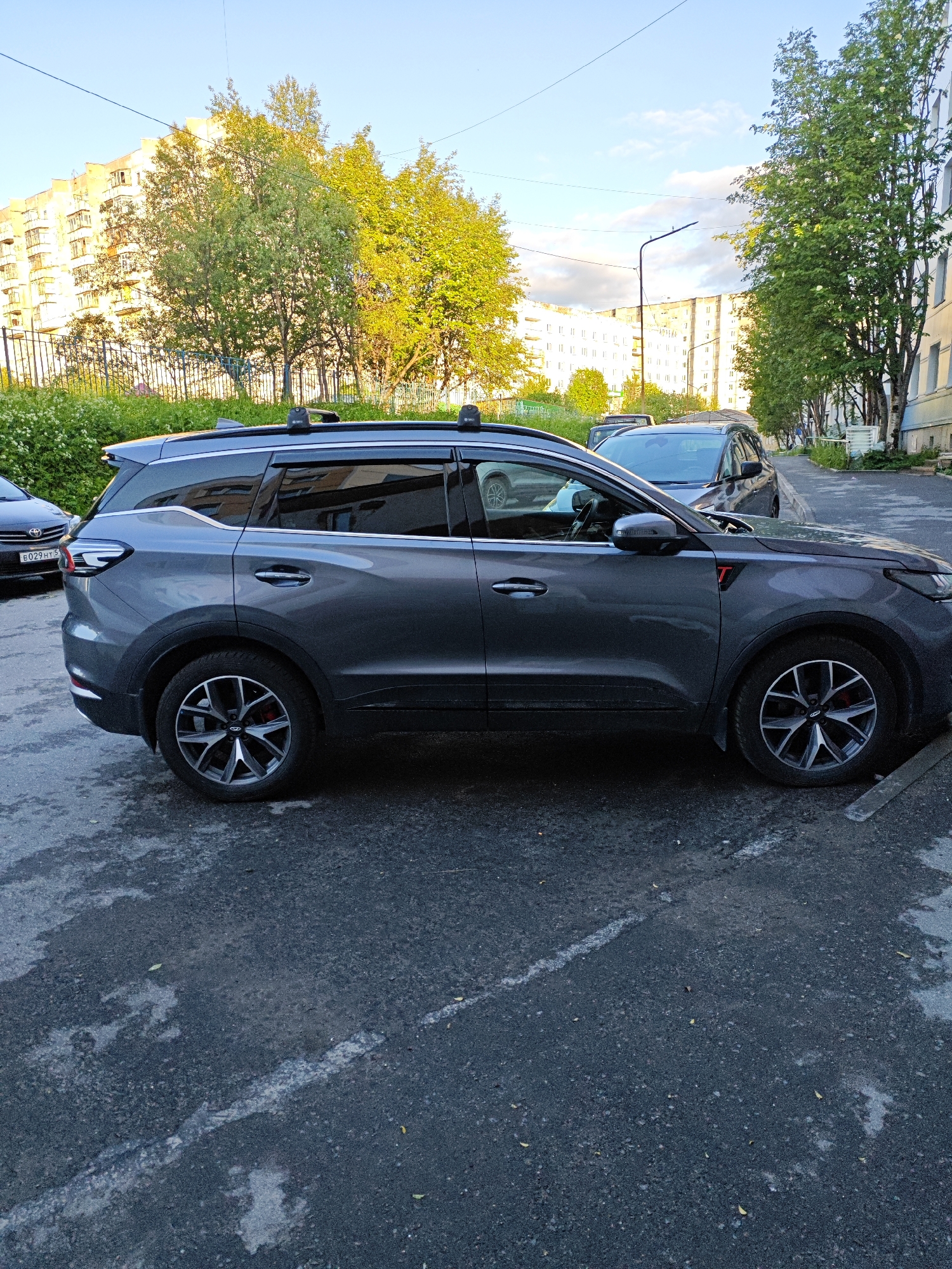 The image depicts a gray SUV parked on an asphalt surface in the city of Polyarny, Russia. The SUV is positioned on the right side of the frame, with its side profile clearly visible. It has a sleek design with tinted windows and roof rails. The vehicle's wheels are a prominent feature, with a multi-spoke design.
In the background, there are apartment buildings typical of Soviet-era architecture, painted in light colors and featuring numerous windows and balconies. Lush green trees with bright green foliage are scattered throughout the scene, some of them illuminated by the soft light of what appears to be late afternoon or early evening sun. A street lamp is visible between the buildings and trees, suggesting an urban street or parking area. Another car, a darker colored SUV, is partially visible in front of the main subject. The asphalt surface shows signs of wear and tear, with some patches of discoloration and a white line painted on it, likely indicating a parking space. The sky is a clear blue with a few wispy clouds. There are no people visible in the image.