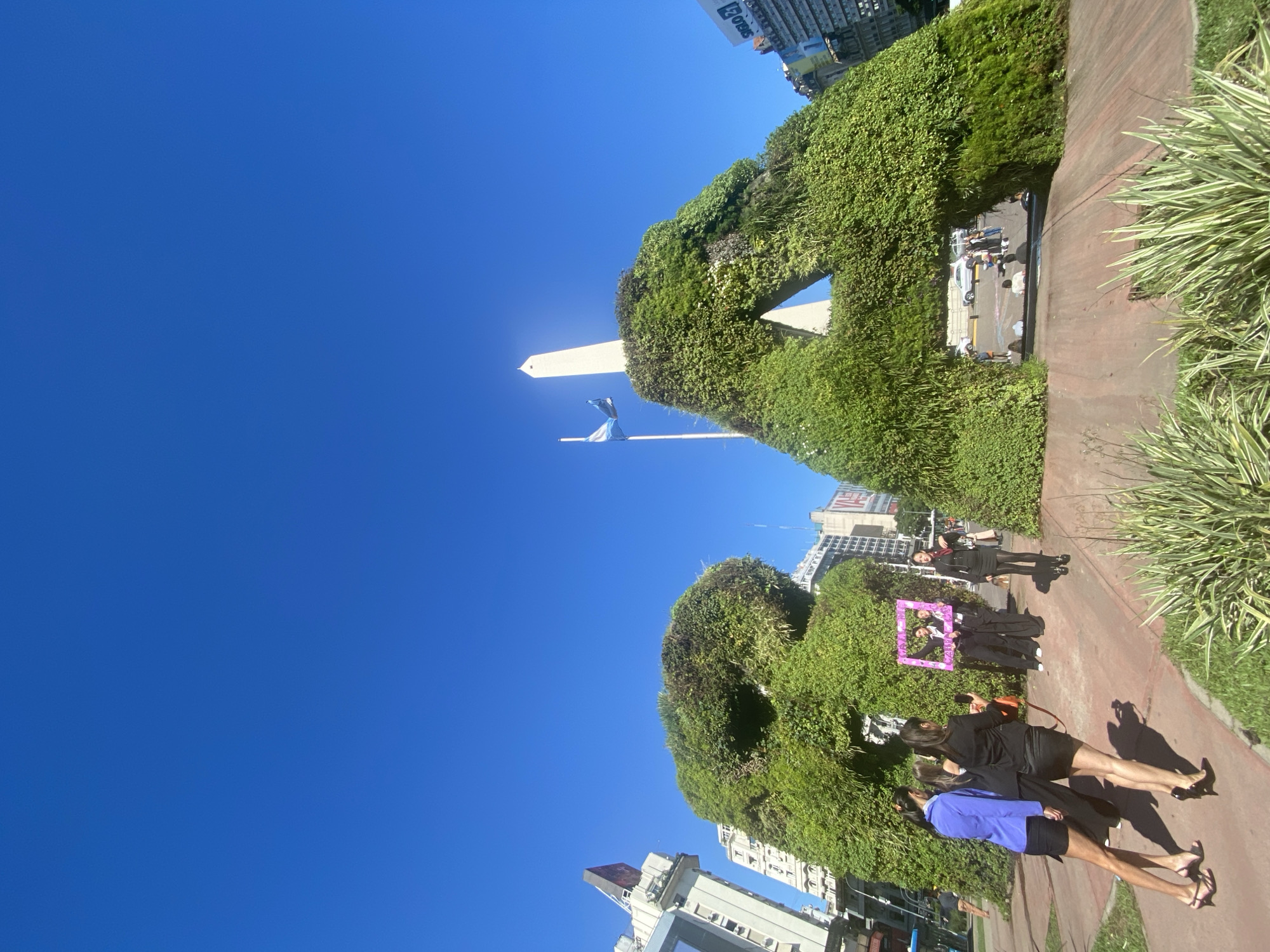 Captured on a clear, sunny day with a brilliant blue sky, this image presents a bustling urban landscape centered around iconic landmarks in Buenos Aires, Argentina. The prominent Obelisco de Buenos Aires, a tall white monument, stands in the mid-ground, accompanied by a flagpole flying the national flag.
The foreground and mid-ground are dominated by expansive, intricately sculpted green hedges or living walls, creating organic, undulating forms that frame the scene. One section of the greenery features a reflective panel or an architectural opening. A reddish-brown paved path runs along the right side, bordered by spiky, variegated plants.
Several people are visible, engaging in various activities. In the foreground, two women walk away from the viewer on the path; one wears a blue top and black shorts/skirt, while the other is dressed in all black. Further down the path, a small group has gathered, with some individuals holding a bright pink/purple empty frame, suggesting a photo opportunity or artistic display. One woman from this group, dressed in black, looks towards the camera. In the background, city buildings are visible, with partial text "F 24/7" and "Obers" (or similar) discernible on one structure.
The strong sunlight creates clear shadows, indicating a midday or early afternoon setting. The overall atmosphere is relaxed, with people enjoying the pleasant weather in what appears to be a public park or plaza.