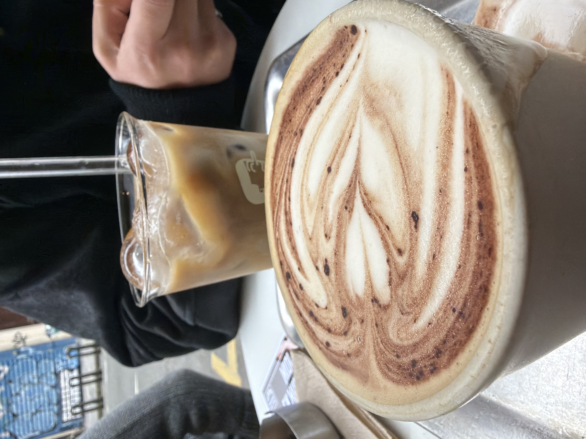 The image depicts a cafe scene, likely indoors. A large mug of cappuccino with latte art in a leaf-like pattern is prominently featured, with a rim of thick foam and brown swirls. Next to it is a glass of iced coffee with a clear straw, a portion of someone's hand resting near the glass. The person is wearing dark clothing. Hints of a white table and a metal tray or coaster are visible under the drinks. In the background, the edge of a blue and white metal chair or railing can be seen, suggesting an outdoor seating area. Considering the style of the cafe and the general aesthetic, this image could plausibly be taken in Paris, France, but without further context, the location remains unconfirmed.