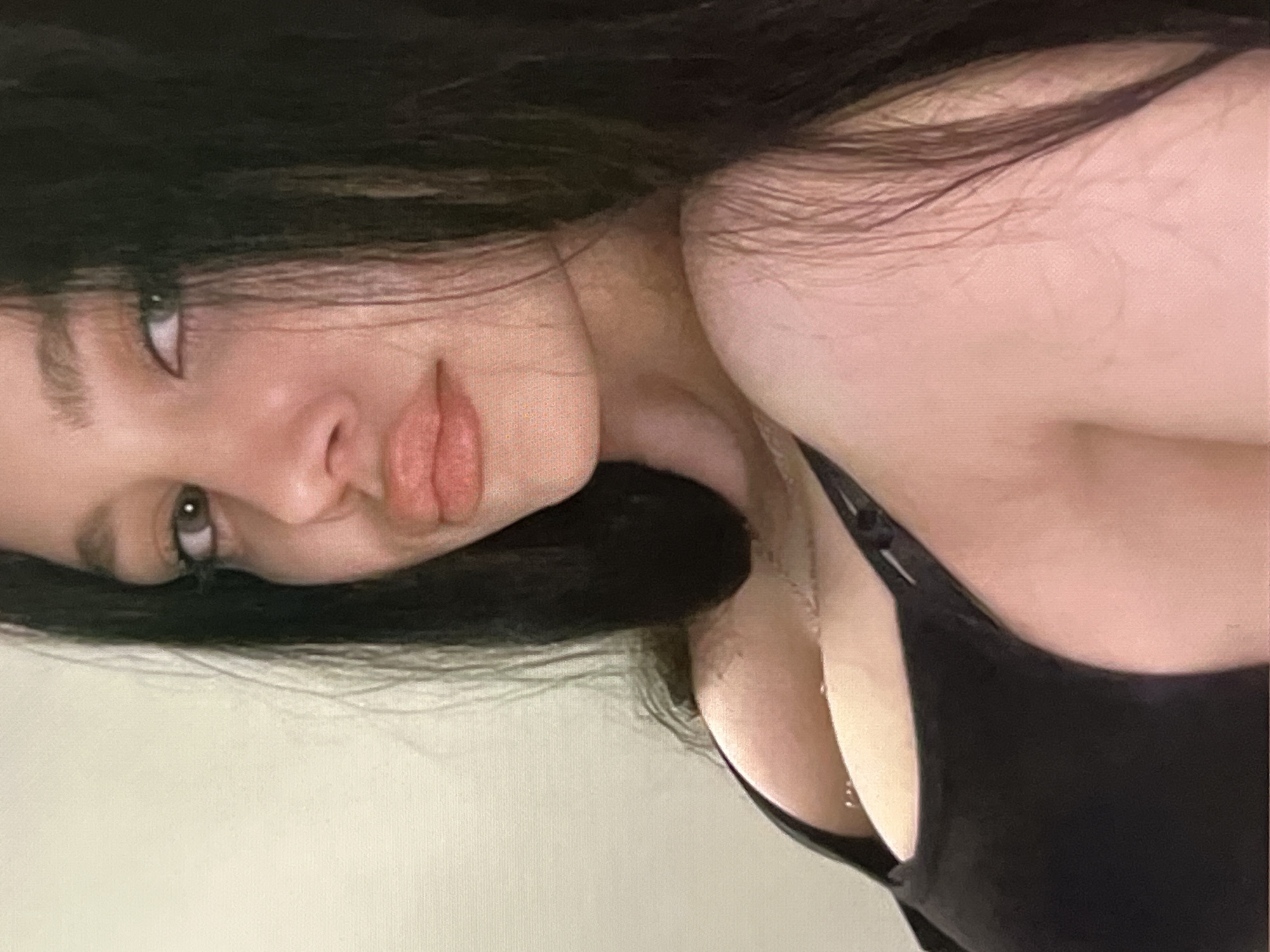 A close-up, medium shot, head and shoulders portrait of a young woman with dark hair, fair skin, and light-colored eyes. She is looking slightly to her left, with her mouth slightly open and her lips appearing full and lightly colored. She is wearing a black tank top, and a thin necklace is visible. The background is a plain, light-colored wall.