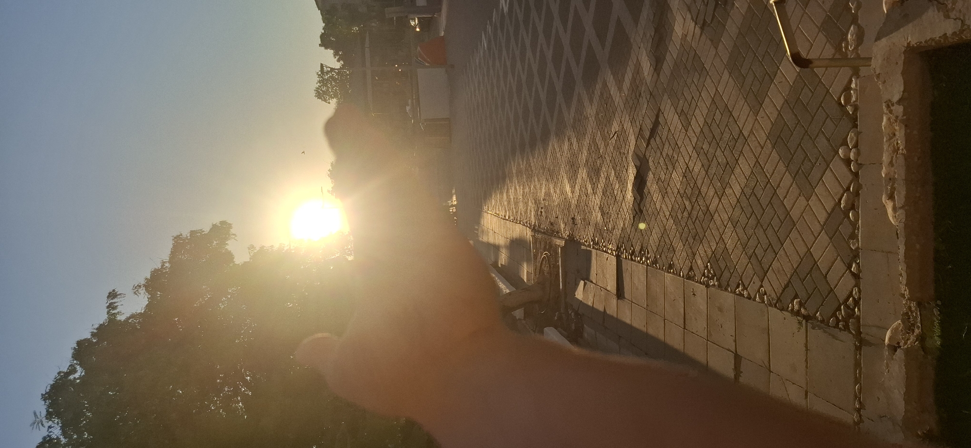 The image captures a sun-drenched scene in Yerocham, Israel, likely during late afternoon or early evening. A bright sun is positioned in the upper center of the frame, partially obscured by a hand and finger in the foreground which is out of focus. The sun's rays create a lens flare effect, casting a warm glow over the entire image.
To the left, the silhouette of a leafy tree is visible against the bright sky. The ground is paved with interlocking rectangular tiles, laid in a herringbone pattern, extending into the distance. A narrow strip of smaller, rounded stones runs along the edge of the paved area, separating it from a tiled walkway and a grassy area in the very bottom of the frame.
In the middle ground, a paved path is visible with a checkered pattern formed by the light and shadows. Further back, there are hints of urban infrastructure, including what appears to be a street lamp and some low-lying structures or stalls, possibly in a park or public square. A small bird is captured mid-flight in the sky, near the sun. The overall atmosphere is one of serene daylight, with the setting or rising sun dominating the visual experience.