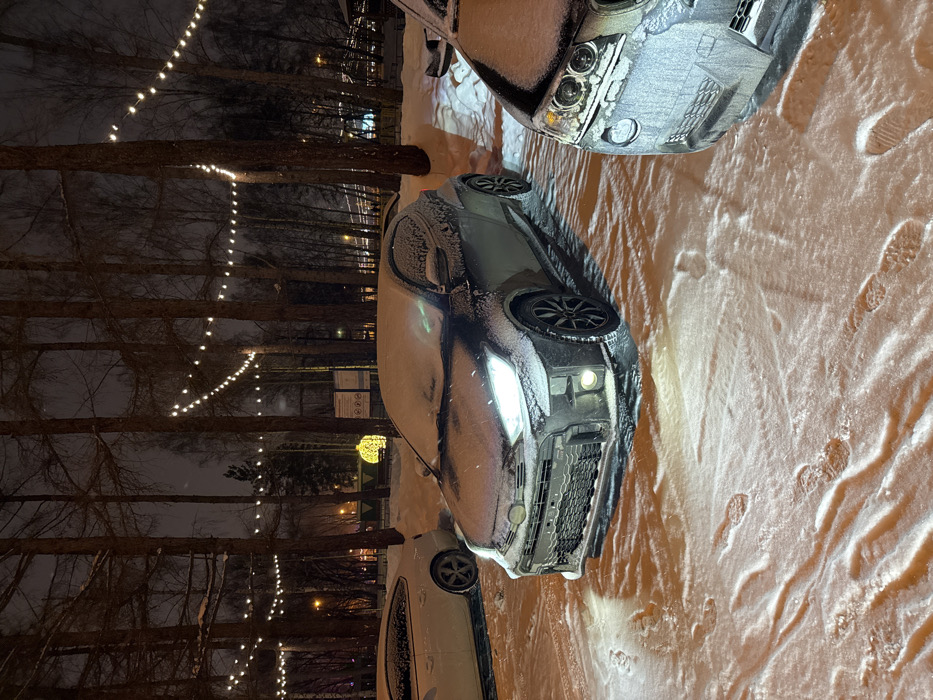 The image displays a winter night scene in Yekaterinburg, Russia, likely within Mayakovsky Central Park of Culture and Leisure. The ground and all visible surfaces are covered in fresh snow. Numerous tall, bare deciduous trees line the area, with decorative string lights suspended horizontally between their trunks, providing ambient illumination.
In the foreground and center, a dark gray Toyota GR Yaris is parked, its body and roof lightly coated with snow. Its headlights and fog lights are illuminated, casting bright cones onto the snowy ground directly in front of the vehicle. To the immediate right, the front-right quarter of a dark, snow-covered SUV or large sedan is partially visible. Further in the background, to the left, the rear-left section of a white, snow-covered coupe is also parked.
The snow-covered ground in the immediate foreground exhibits multiple clear footprints, indicating recent pedestrian activity. In the mid-background, visible through the trees, a large, spherical yellow light fixture serves as a decorative element. A signpost with unreadable text is also visible in this area. Distant city lights and additional trees are discernible in the far background. No people are visible in the scene.