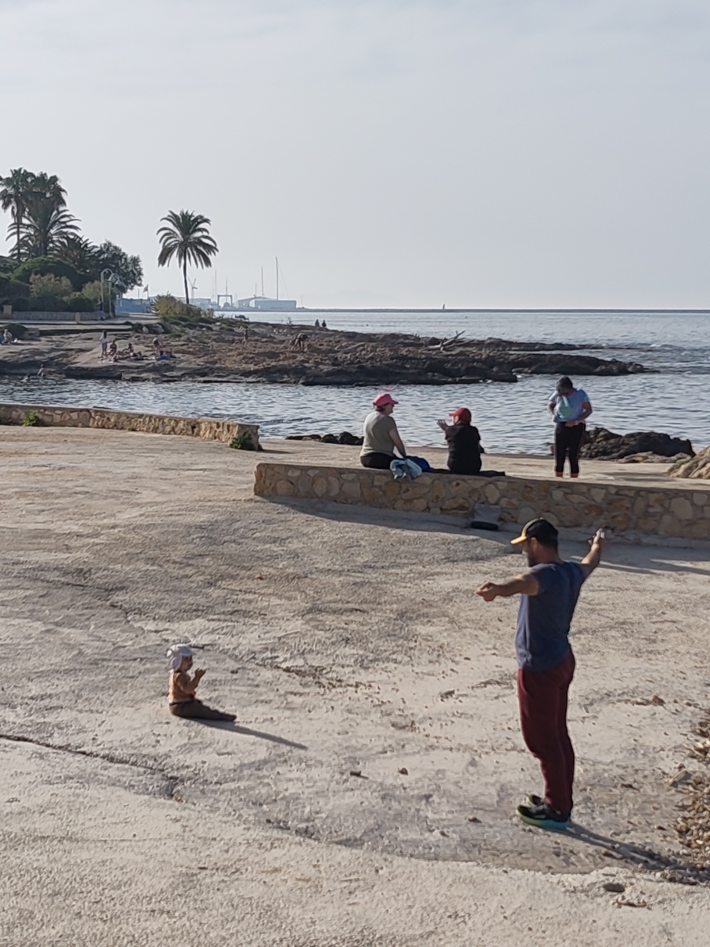 A concrete boardwalk in Rotes, Spain, is seen with a group of people. A man in a blue t-shirt and maroon pants with a cap is standing on the right, arms outstretched. In the foreground a baby or small child sits on the boardwalk. Further away, three women are sitting on the edge of the boardwalk, with one standing in the water. People are visible on the beach. Palm trees are visible and other vegetation near the beach. There are some boats on the horizon.