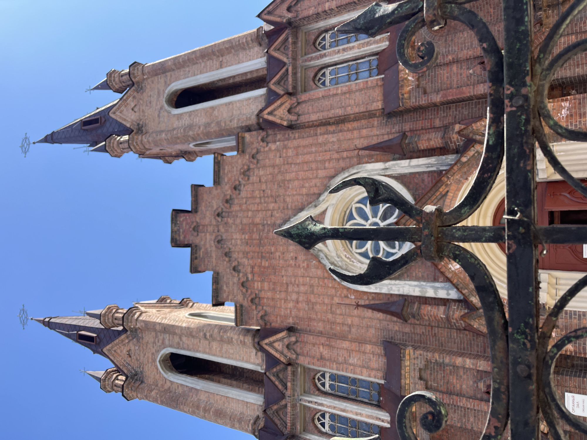 A low-angle, vertically-oriented photograph shows the top portion of the Roman Catholic Church of the Transfiguration in Krasnoyarsk, Russia, against a clear blue sky.
The church is constructed from red brick and features gothic architectural elements. The main body of the church visible in the frame is flanked by two towers, each with a pointed spire. The towers feature arched windows and decorative detailing.
In the foreground, an ornate metal fence partially obscures the church's facade. The fence is painted black and shows signs of age and wear. It has intricate, swirling designs.
The photograph is taken from a position that looks upwards, emphasizing the height of the church towers and the ornate fence.