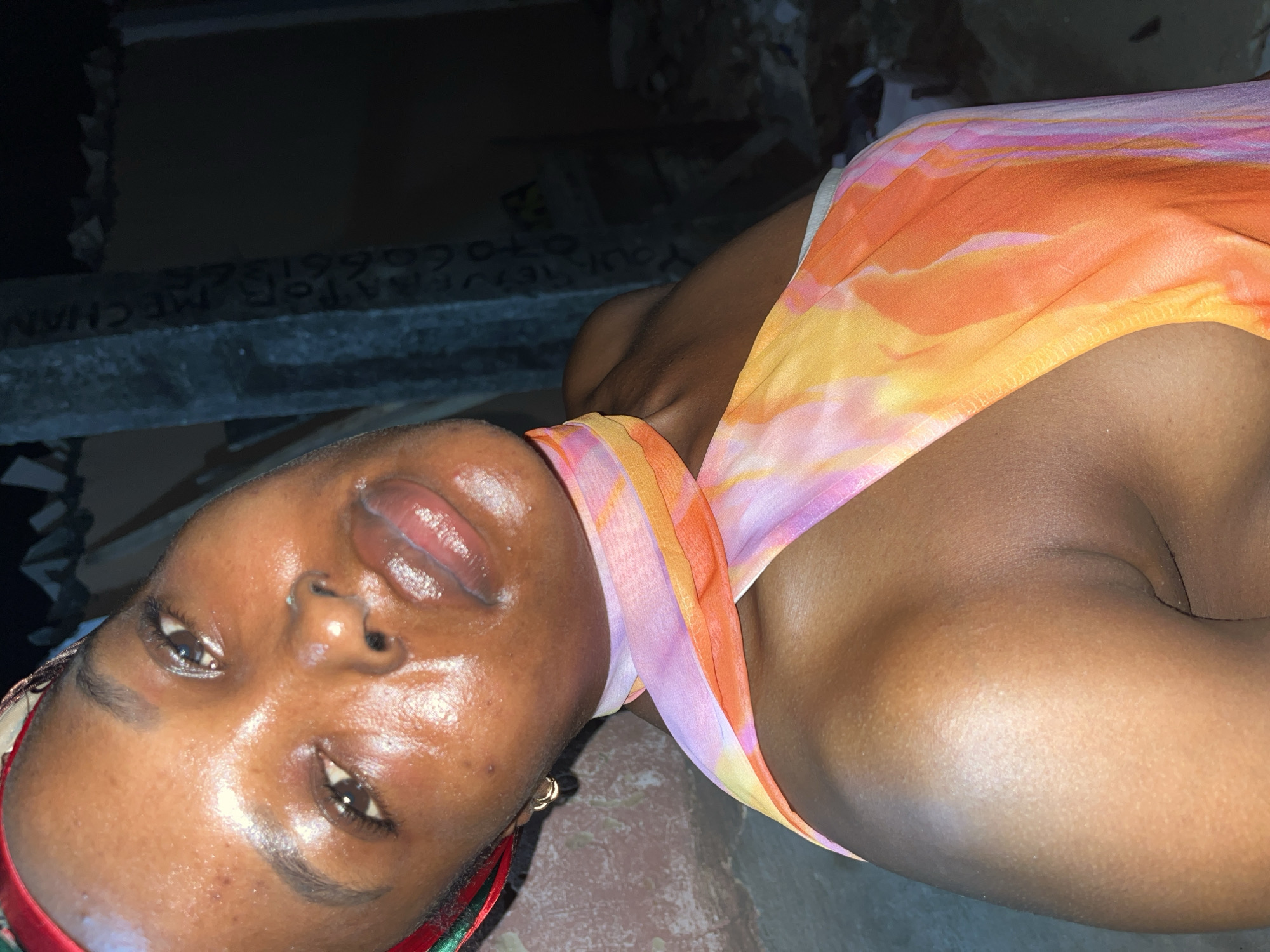 A close-up, rotated image depicts a woman with dark skin. Her face is shiny with moisture, and she has full, glossy lips. She is wearing a colorful top with a blend of orange, yellow, and light purple hues. A red and green patterned headscarf is visible at the bottom left corner. The background is dark and indistinct, but a partial view shows a concrete structure with faded white lettering. Based on the lettering "ATOR, MECHAN...070661326" and the implied subject, this image potentially shows a woman in or near a mechanic shop or auto parts store located in Ado, Nigeria.