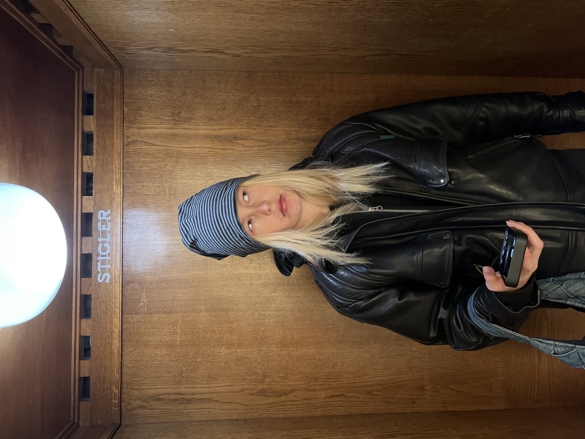 A single individual is observed standing inside a wooden-paneled elevator, indicated by the name "STIGLER" inscribed in silver lettering on the decorative wooden trim above. The presence of a Stigler elevator points to a building located in Strasbourg, France. Overhead, a large, opaque spherical light fixture provides illumination. The subject, appearing to be a young adult with long, straight blonde hair, wears a grey and black striped beanie, a black leather jacket with multiple pockets, and a dark, zippered garment underneath. Their gaze is directed towards the upper right, with a neutral to slightly pensive facial expression and parted lips. In their left hand, they hold a small, black electronic device with a grid of circular depressions on its surface, along with the light blue, quilted fabric strap of a shoulder bag. The immediate environment consists solely of the light brown wooden paneling forming the elevator interior.
