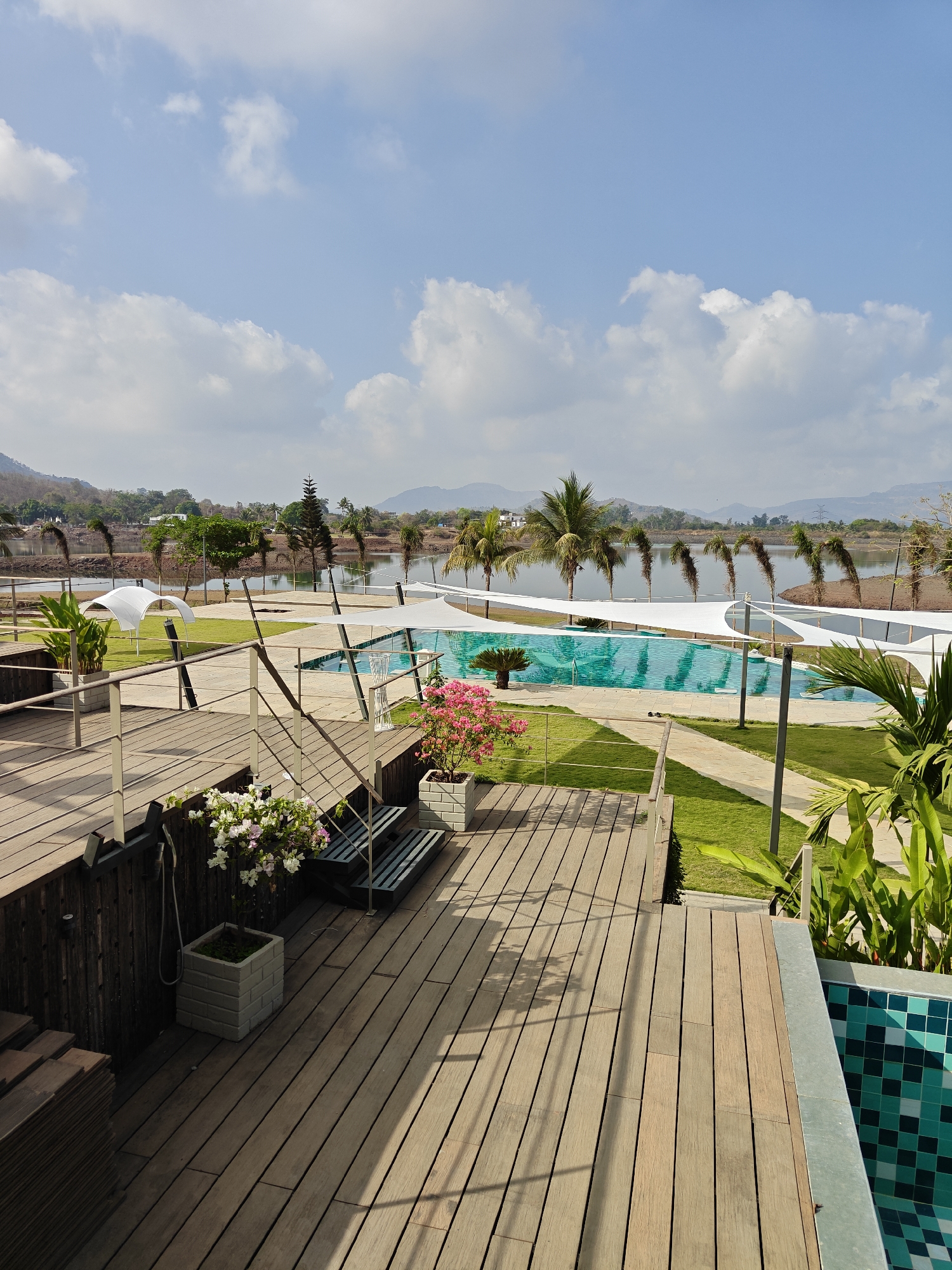 The photograph depicts a serene poolside setting in Kalote Mokashi, India. A wooden deck occupies the foreground, with potted plants adding touches of color. The deck overlooks a turquoise swimming pool, partially shaded by white awnings. Beyond the pool, a calm lake stretches to the horizon, framed by palm trees and distant hills. The sky is blue with scattered white clouds. No people are visible in the scene.